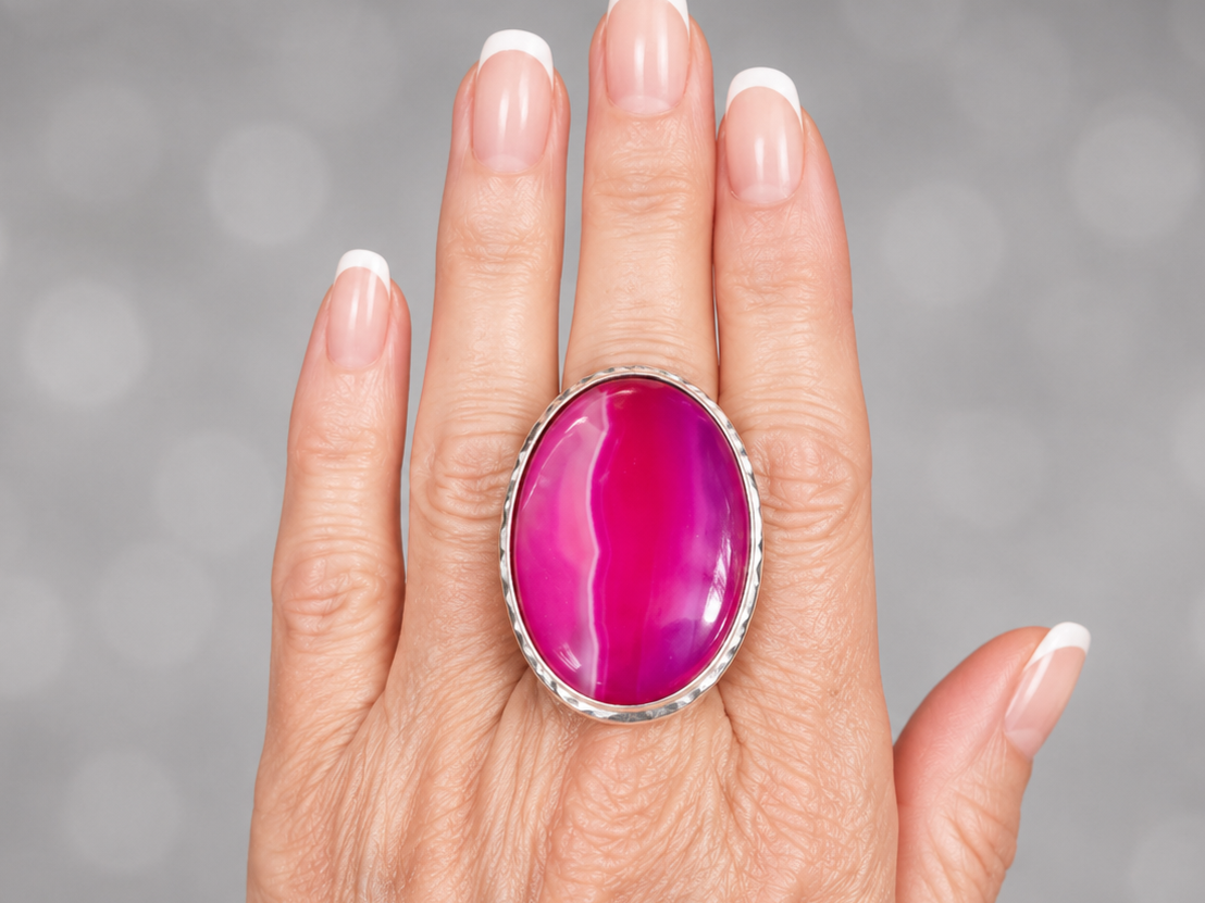 A close-up of a hand wearing a large oval ring with a vivid magenta-pink cabochon stone in a silver-toned bezel setting, against a soft gray bokeh background.