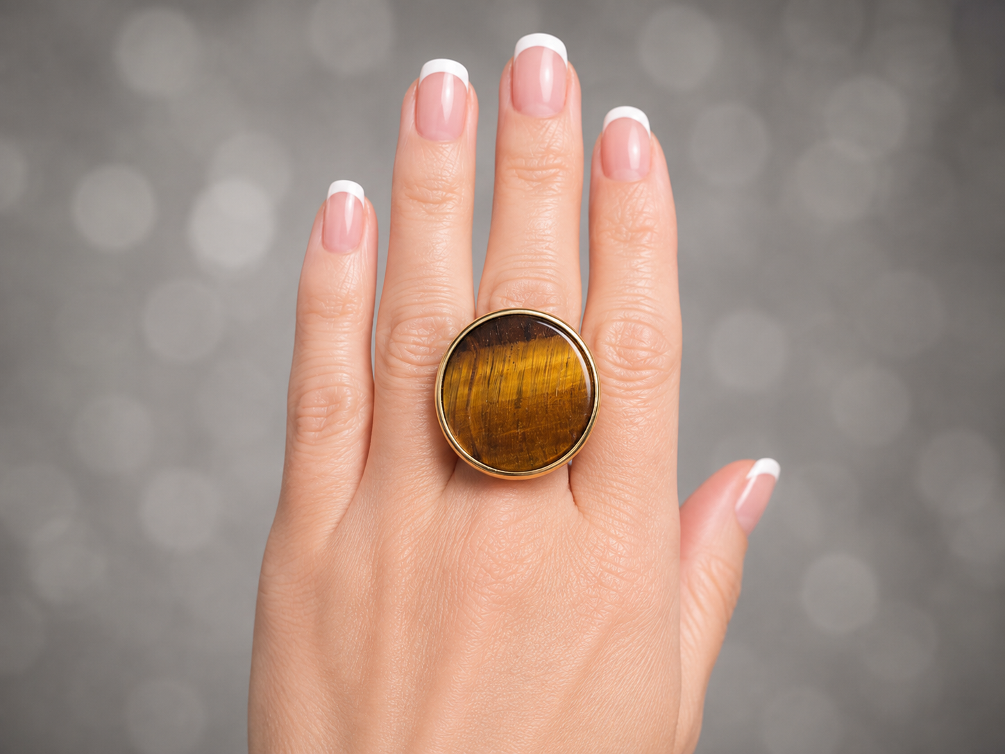 A close-up of a hand with a French manicure wearing a large round ring with a golden-yellow tiger's eye stone in a gold bezel setting, against a soft gray bokeh background.