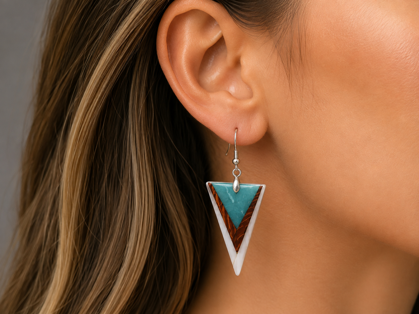 A close-up of a woman's ear wearing a triangular dangle earring with turquoise blue, white, and brown inlay details on a silver hook.