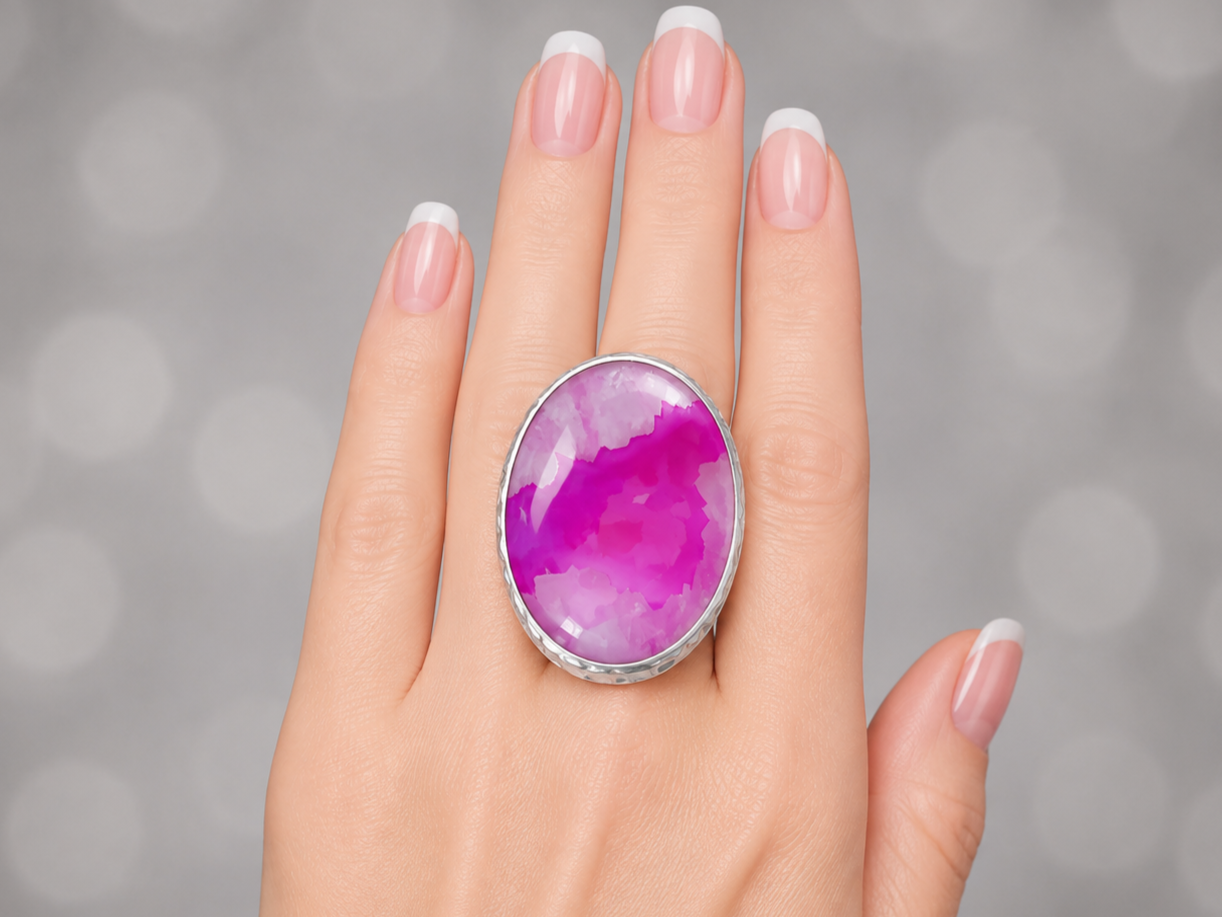 A close-up of a hand with a French manicure wearing a large oval ring featuring a vibrant magenta and purple agate cabochon set in a silver bezel, against a soft gray bokeh background.