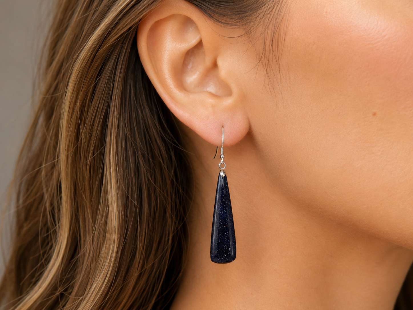 A close-up of a model's ear wearing a dark navy blue teardrop-shaped stone dangle earring with a silver fishhook backing.