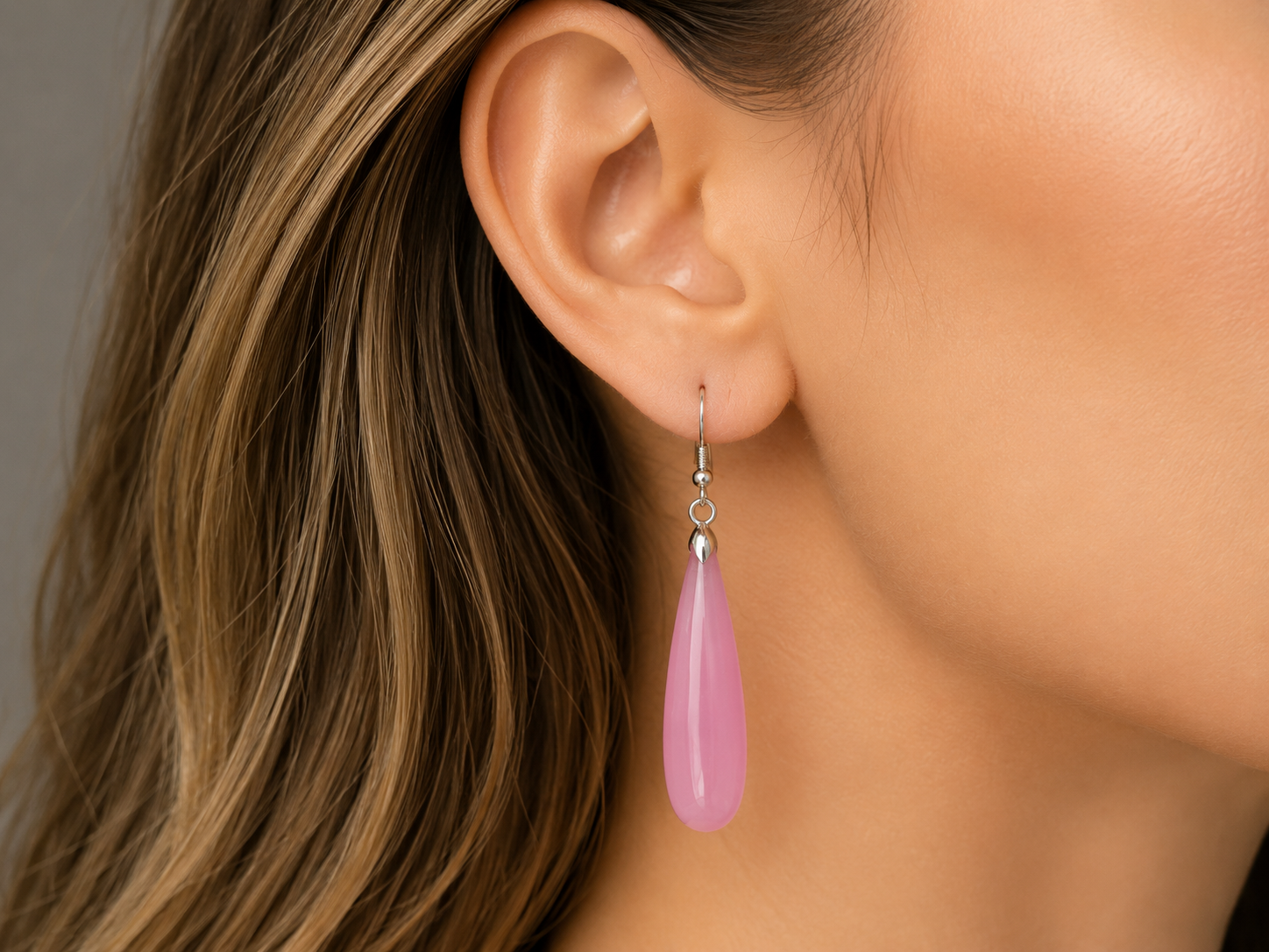 A close-up of a woman's ear wearing a single dangling teardrop-shaped pink stone earring with a silver hook.