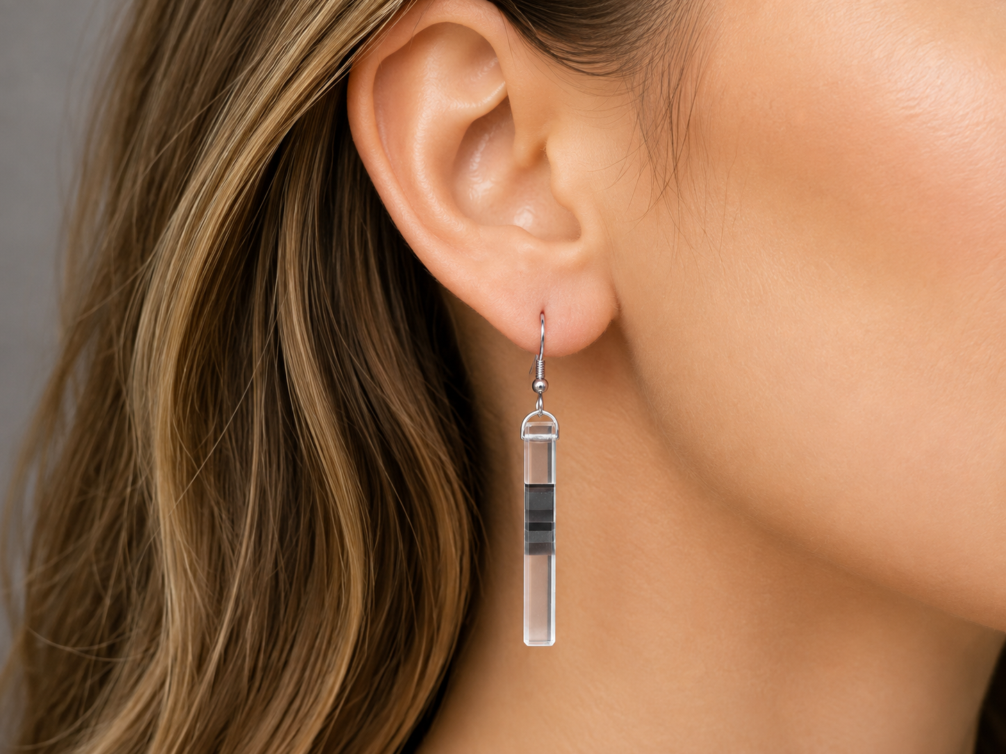 A close-up of a woman's ear wearing a long rectangular drop earring made of clear material with a black horizontal stripe and a silver hook. Main image