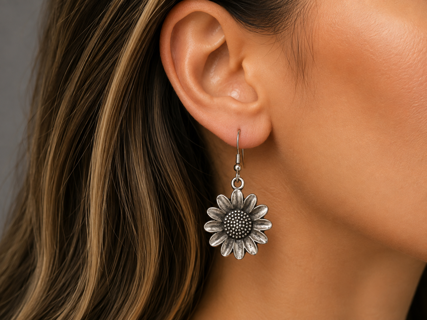 A close-up of a woman's ear wearing a dangling flower-shaped earring in an antiqued silver finish.