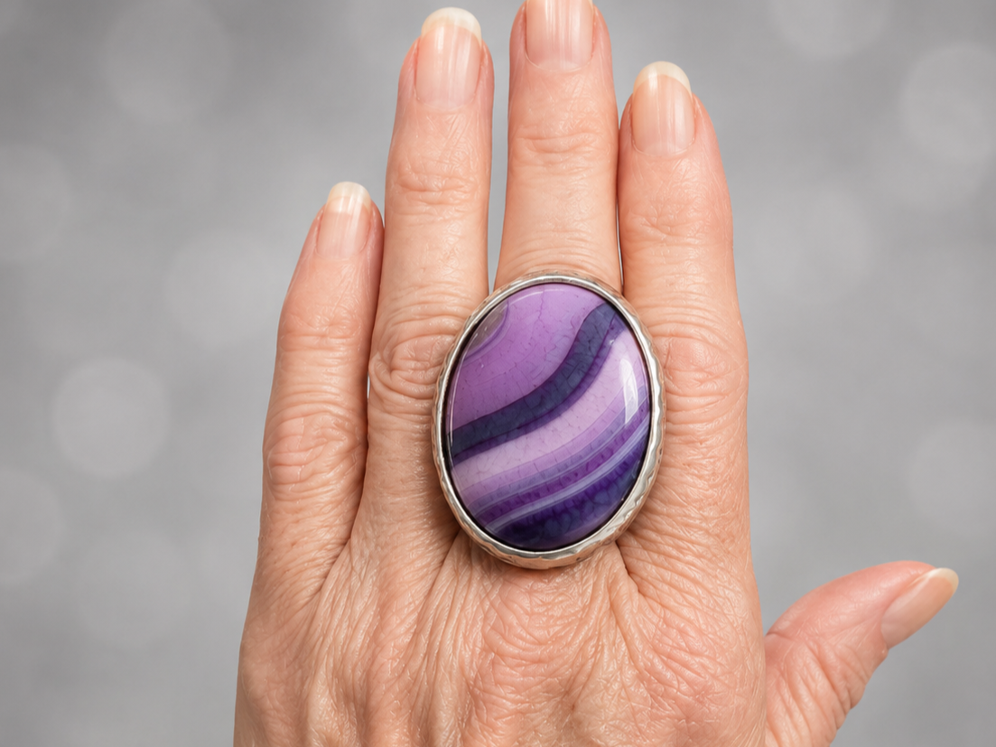 A close-up of a hand wearing a large oval ring with a polished purple and lavender banded stone in a silver bezel setting, against a soft gray bokeh background.