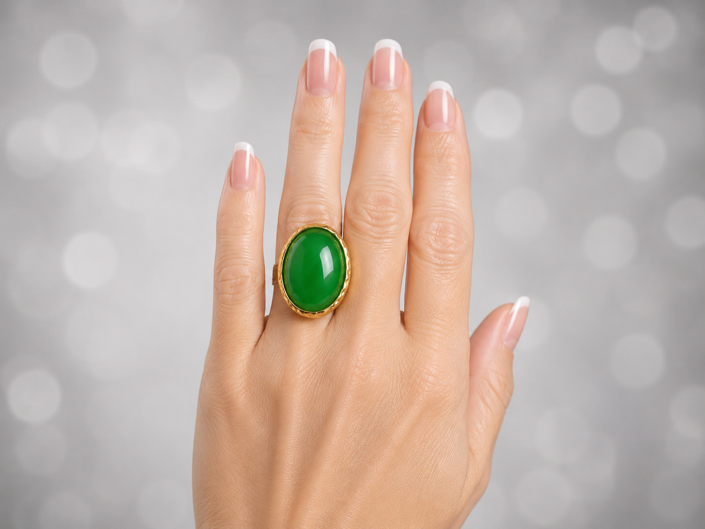 A gold ring with a large oval green stone, worn on a hand with a French manicure, set against a soft gray bokeh background.
