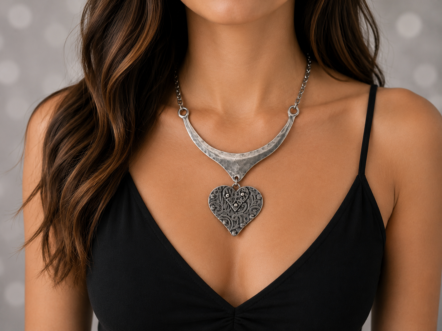 A close-up of a woman wearing a silver crescent-shaped necklace with an ornate heart pendant, styled with a black spaghetti-strap top against a soft gray bokeh background. Main image