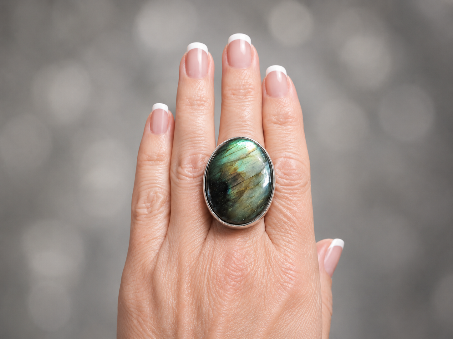 A close-up of a hand with a French manicure wearing a large oval labradorite ring with a silver bezel, set against a soft, blurred gray bokeh background.
