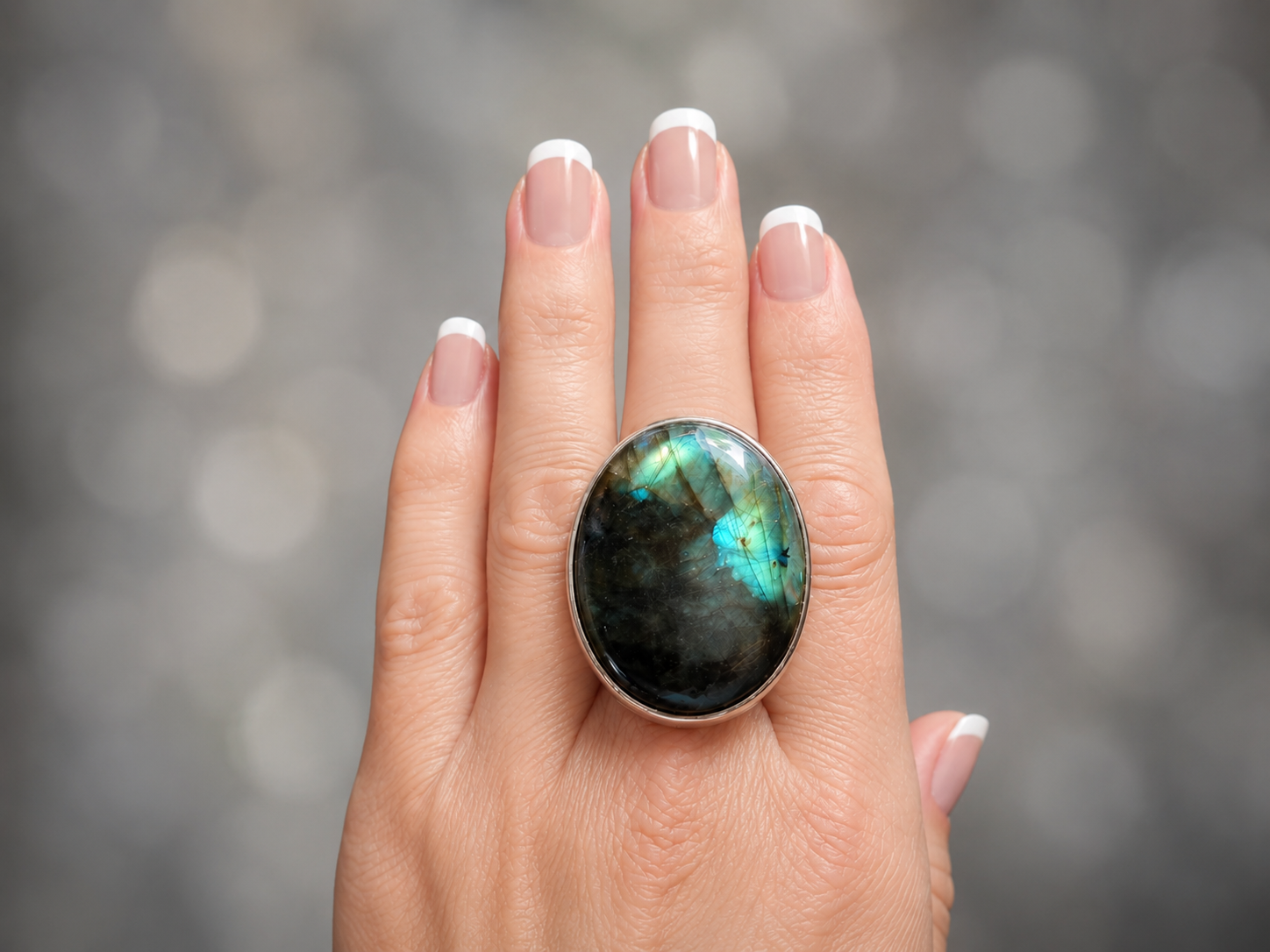 A close-up of a hand wearing a large oval ring featuring a dark labradorite stone with vibrant green and blue iridescent flashes, set in a silver-toned metal bezel.