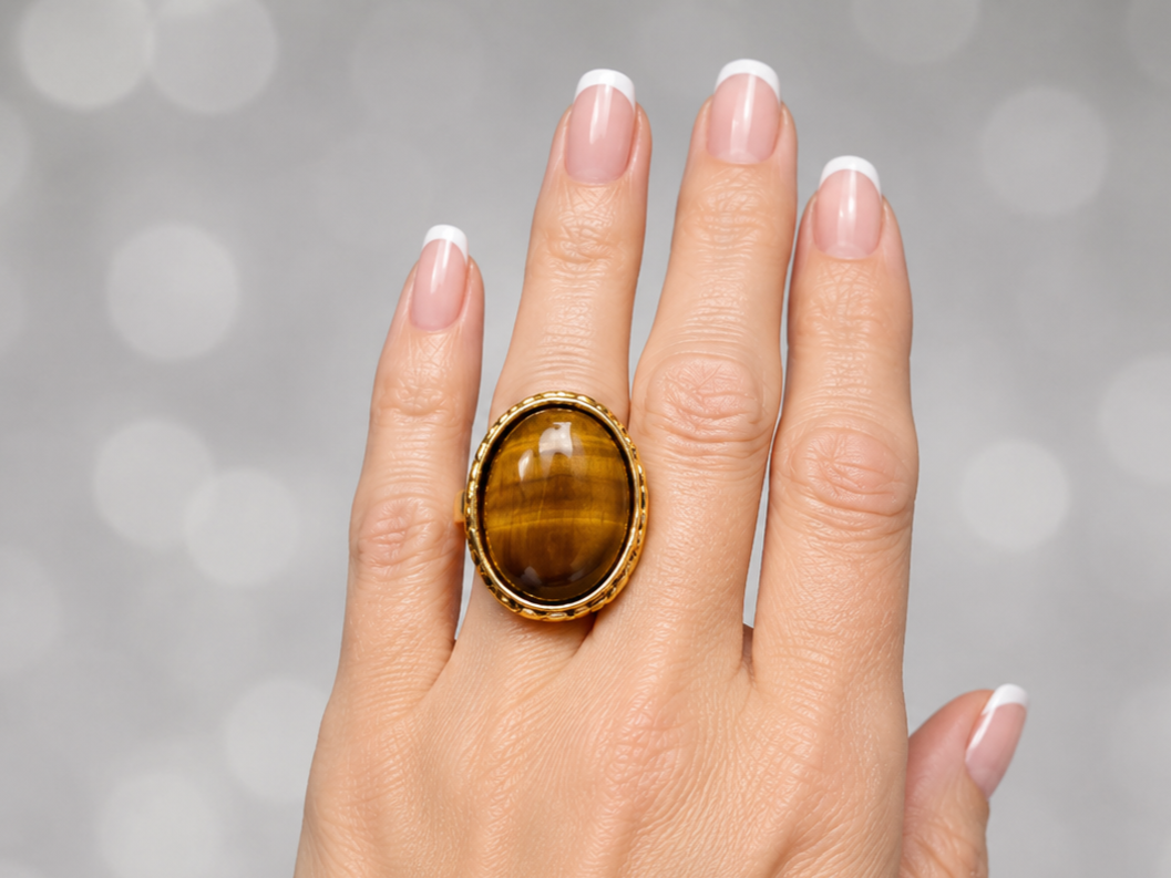 A close-up of a hand wearing a large oval ring with a polished tiger's eye stone in a gold bezel setting, against a soft gray bokeh background.