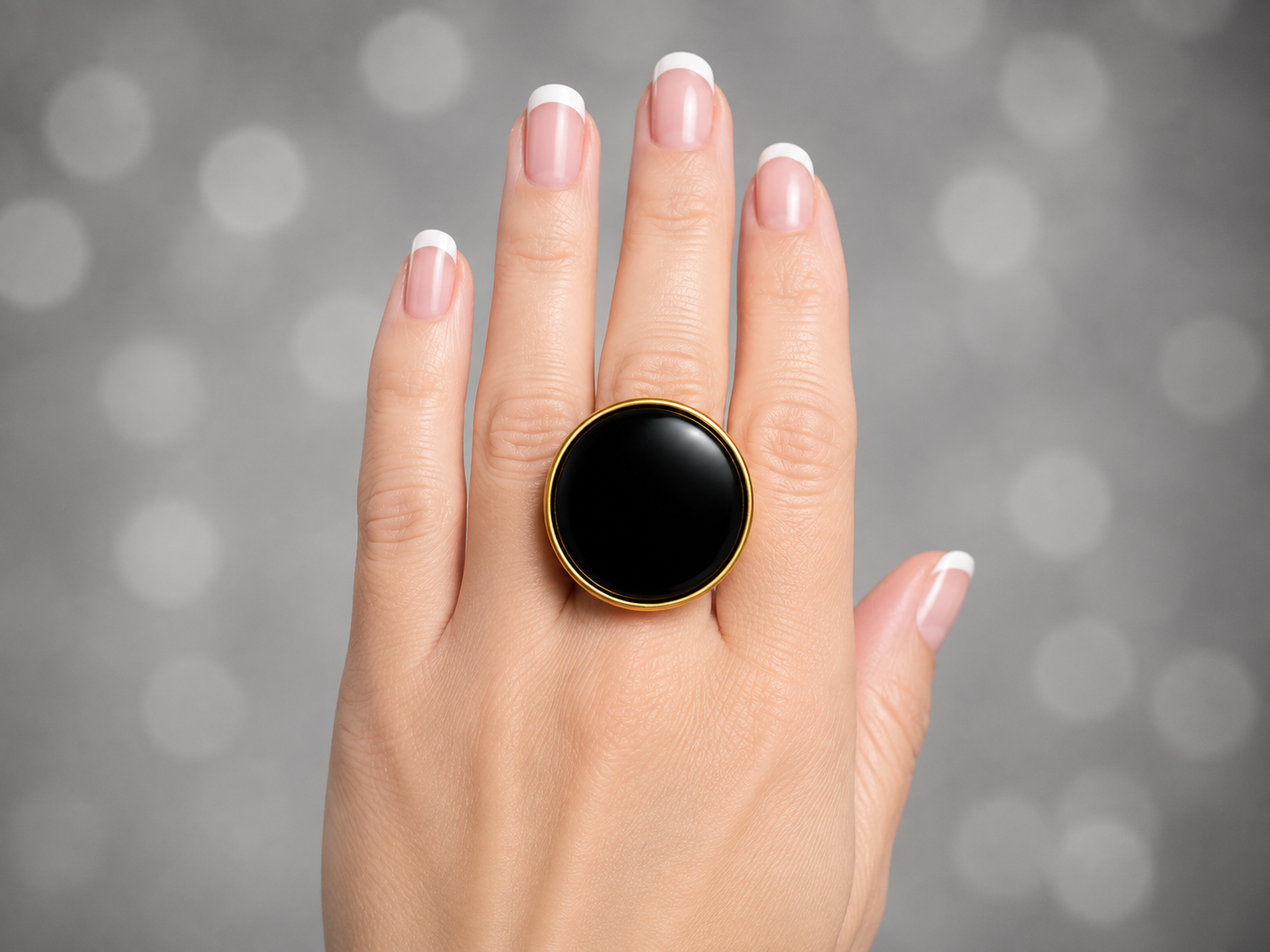 A close-up of a hand with a French manicure wearing a large round black onyx ring with a gold bezel, set against a soft gray bokeh background.