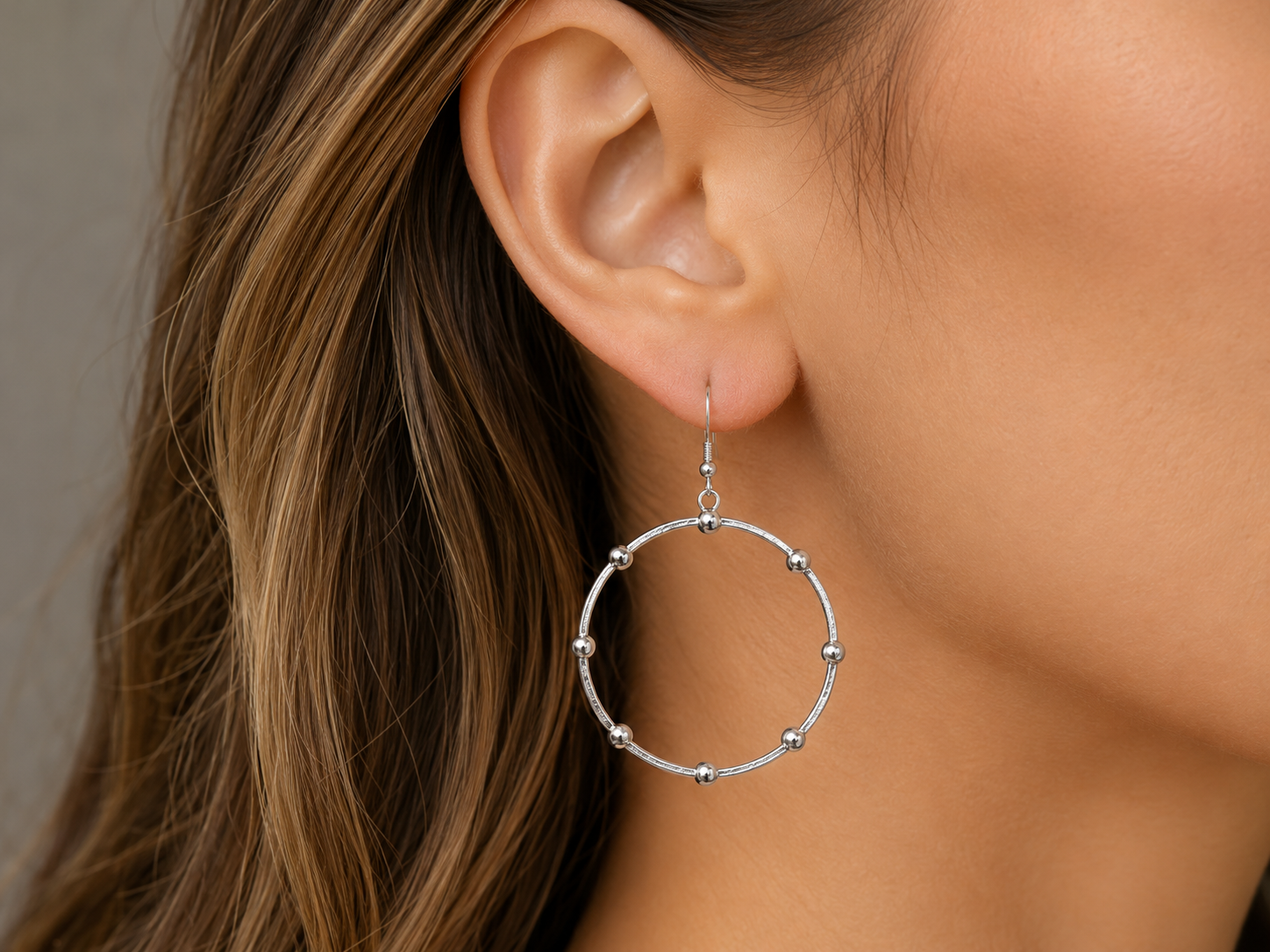 A close-up of a woman's ear wearing a silver hoop earring with small bead accents along the outer edge, against a soft neutral background.