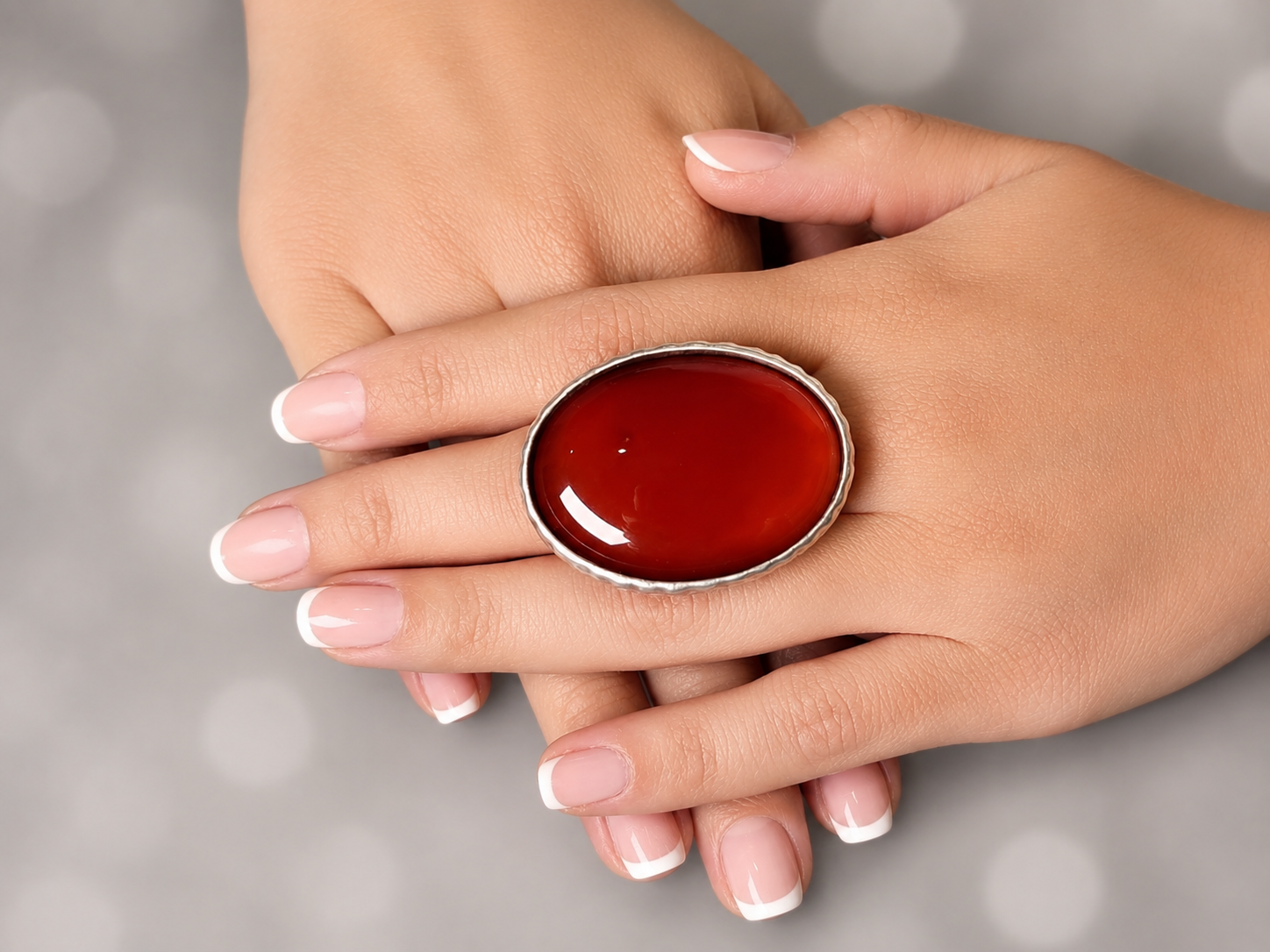 A close-up of hands with a French manicure wearing a large oval silver ring set with a glossy deep red cabochon gemstone, set against a soft gray bokeh background.