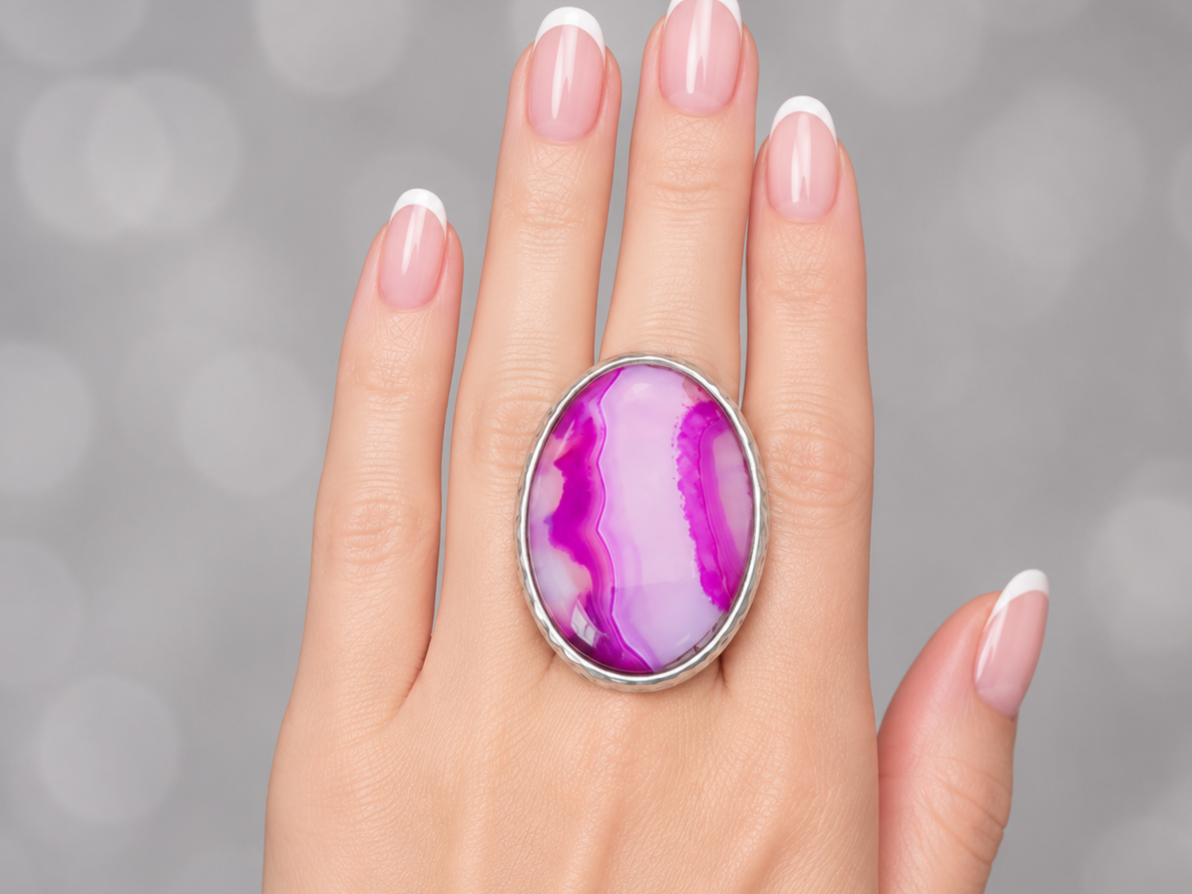 A close-up of a hand with a French manicure wearing a large oval silver ring featuring a vibrant purple and pink agate stone.
