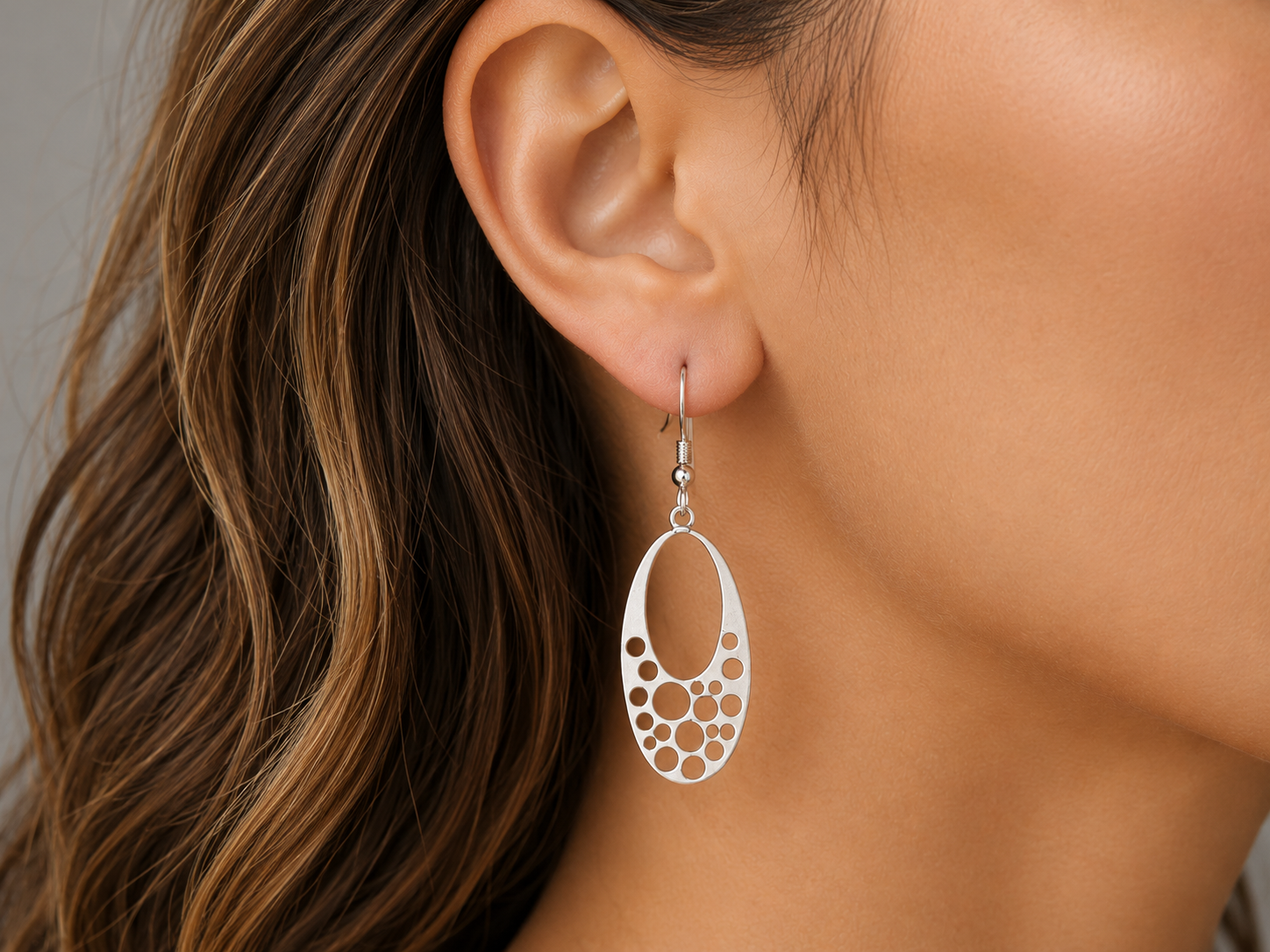 A close-up of a woman's ear wearing a silver dangle earring with an oval cutout design and small circular details.