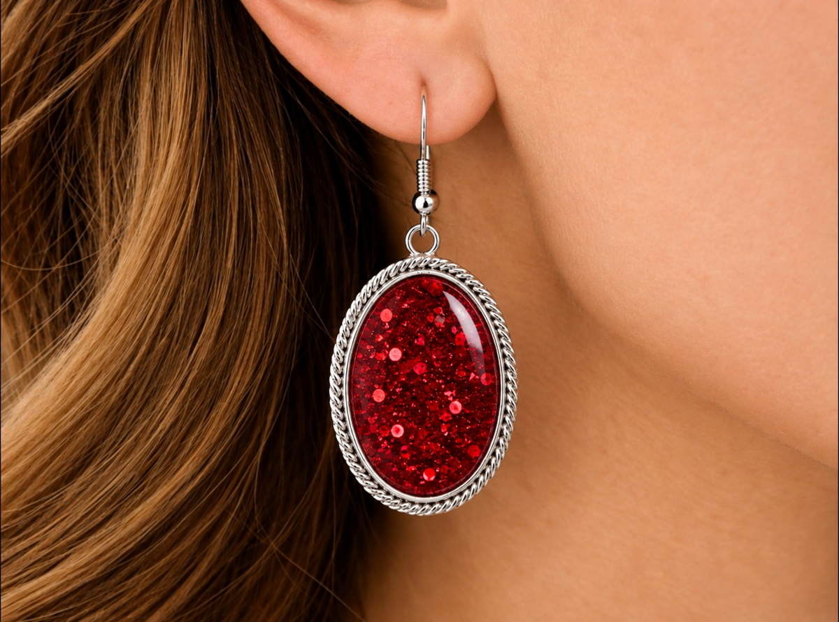 A close-up of a person's ear wearing an oval dangling earring with a silver-toned rope-textured border and a sparkling red center.