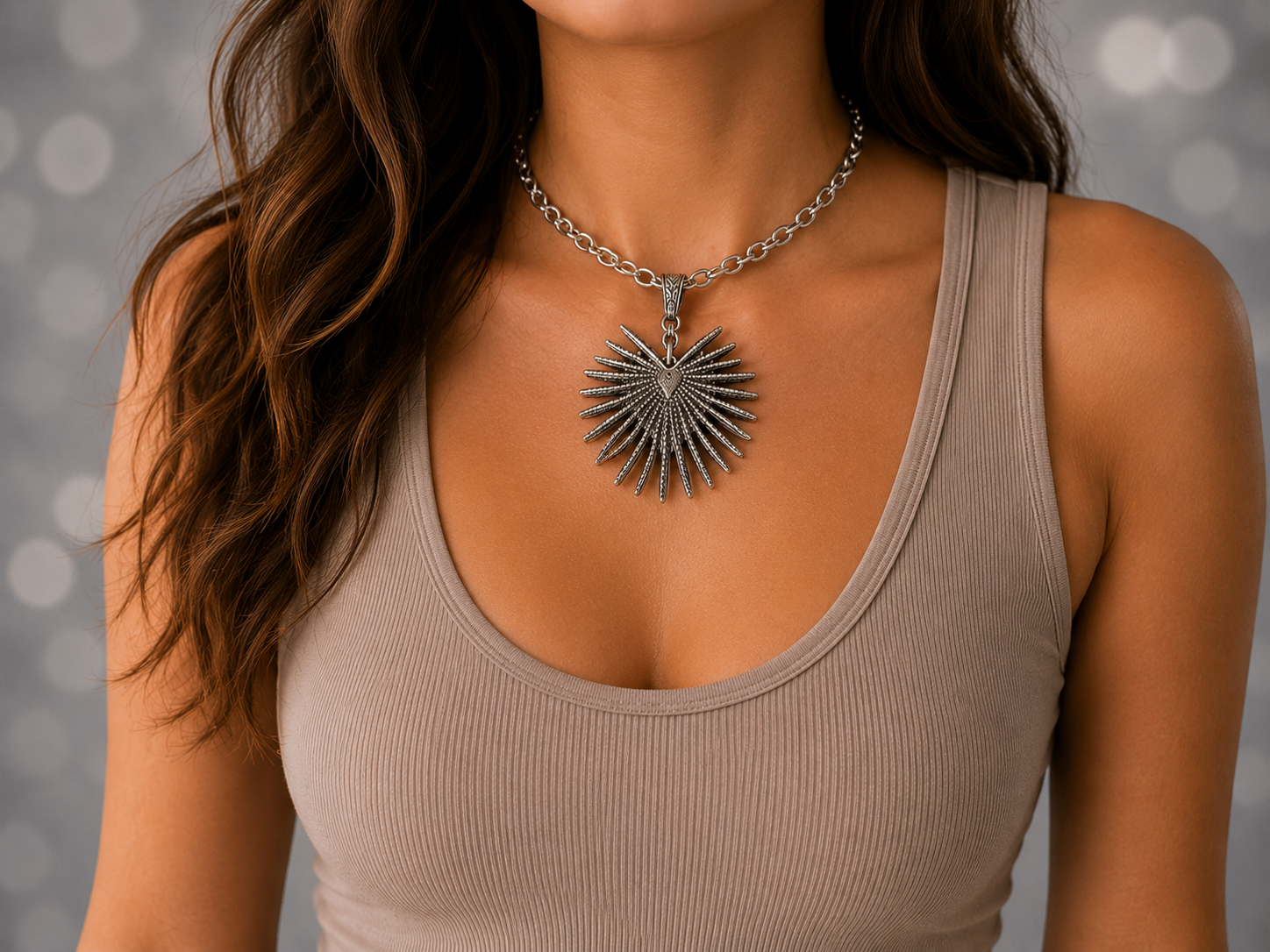 A close-up of a woman wearing a silver sunburst pendant necklace with a chain, paired with a beige ribbed tank top against a soft gray bokeh background.