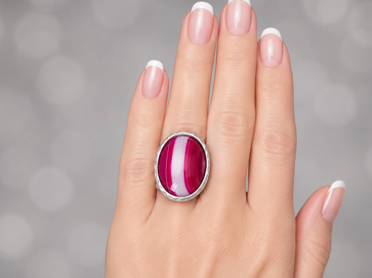 A close-up of a hand with a French manicure wearing a silver ring with a large oval cabochon gemstone featuring pink, magenta, and white horizontal stripes.