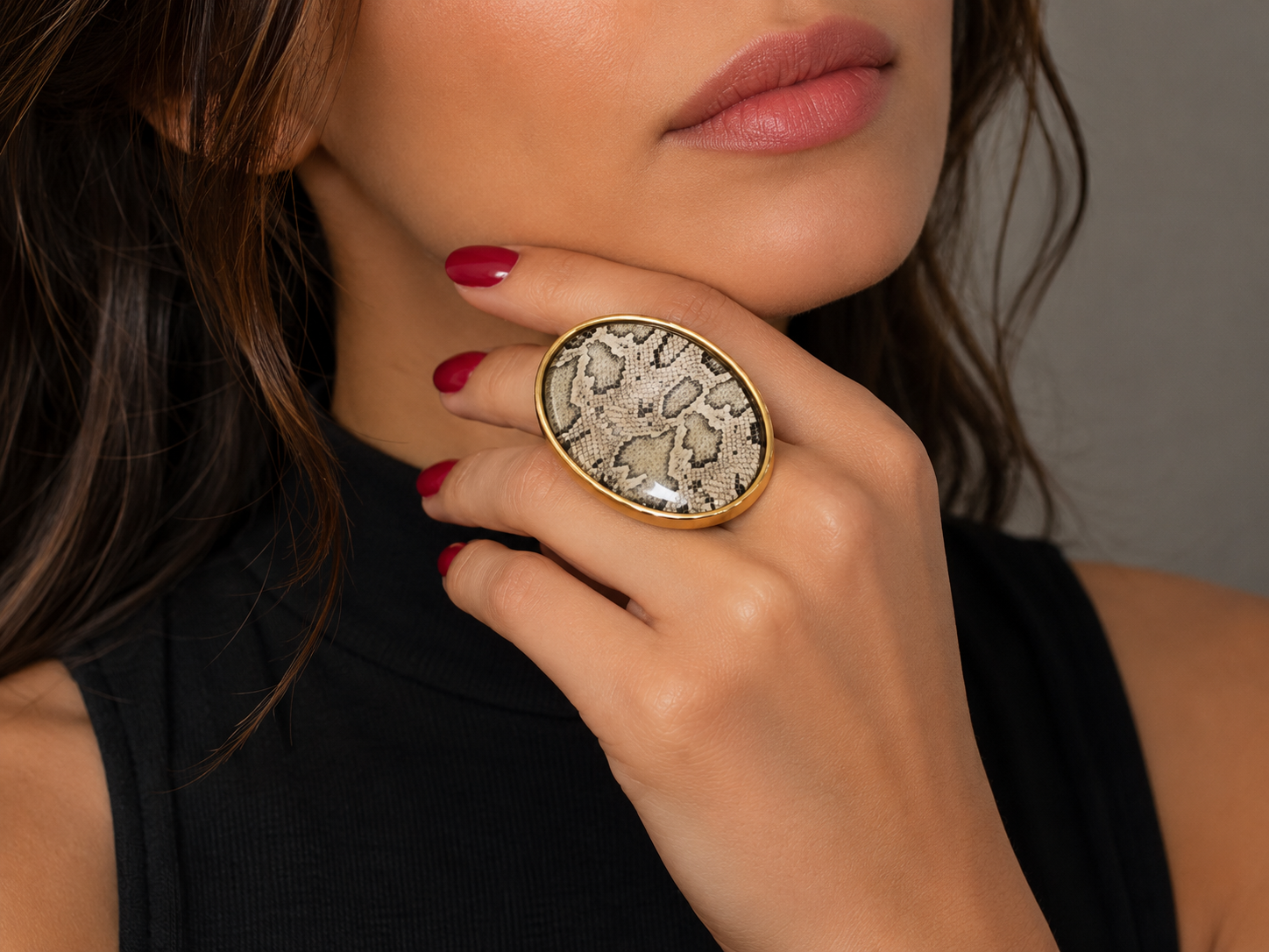 A close-up lifestyle shot of a model wearing a large oval ring with a textured beige and brown snakeskin pattern in a gold-tone frame.