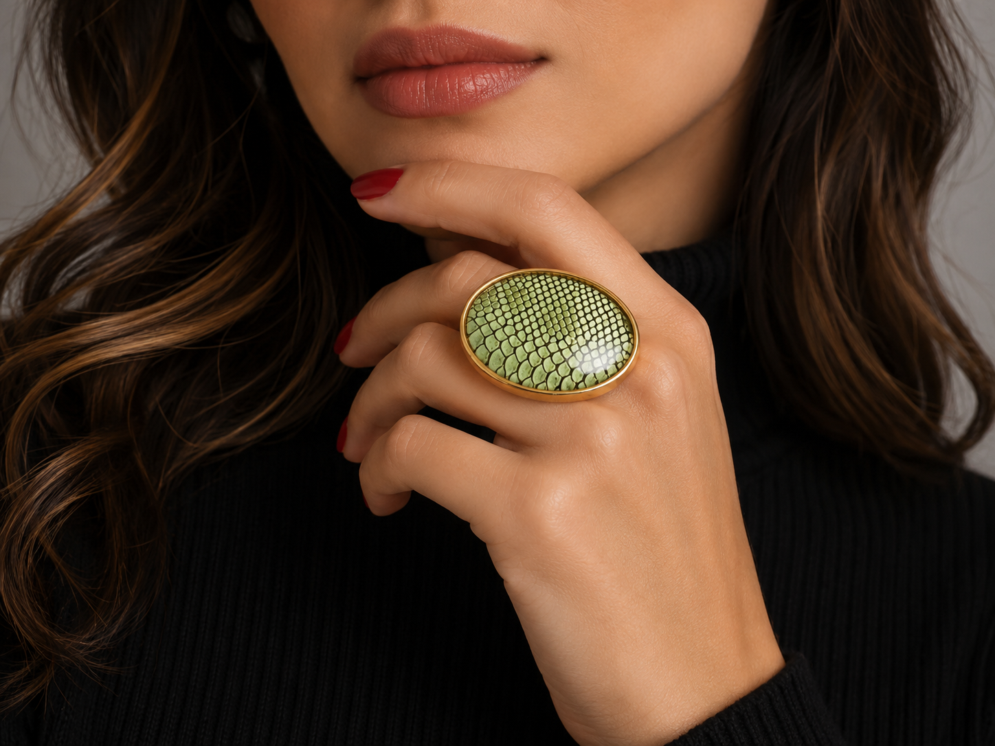 A close-up lifestyle shot of a model wearing a large oval ring with a light green snakeskin pattern and a gold-tone frame, set against a dark ribbed sweater. Main image