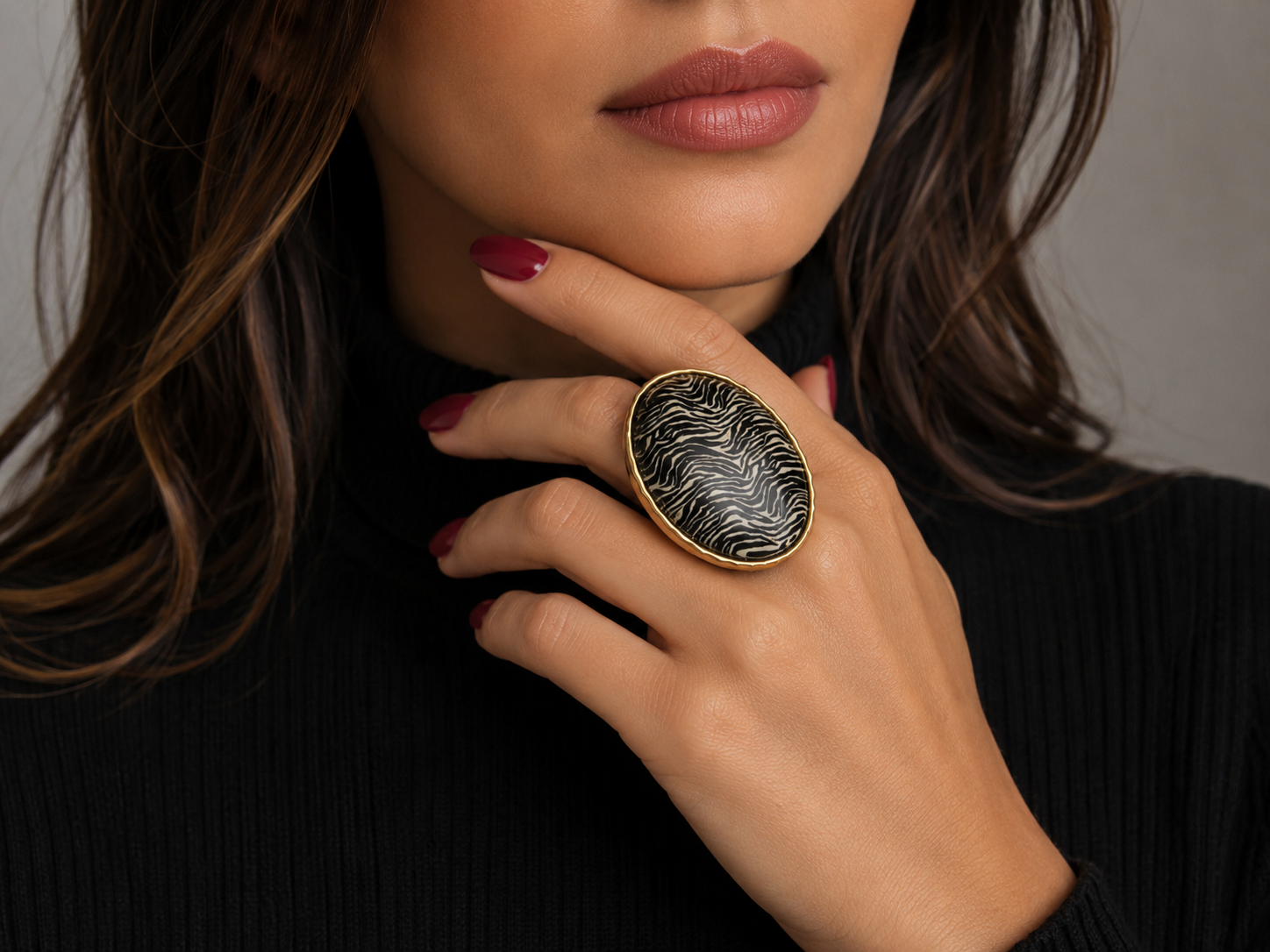 A close-up lifestyle shot of a model wearing a large oval ring with a black and white feather pattern and a gold-toned bezel, set against a dark ribbed turtleneck sweater. Main image