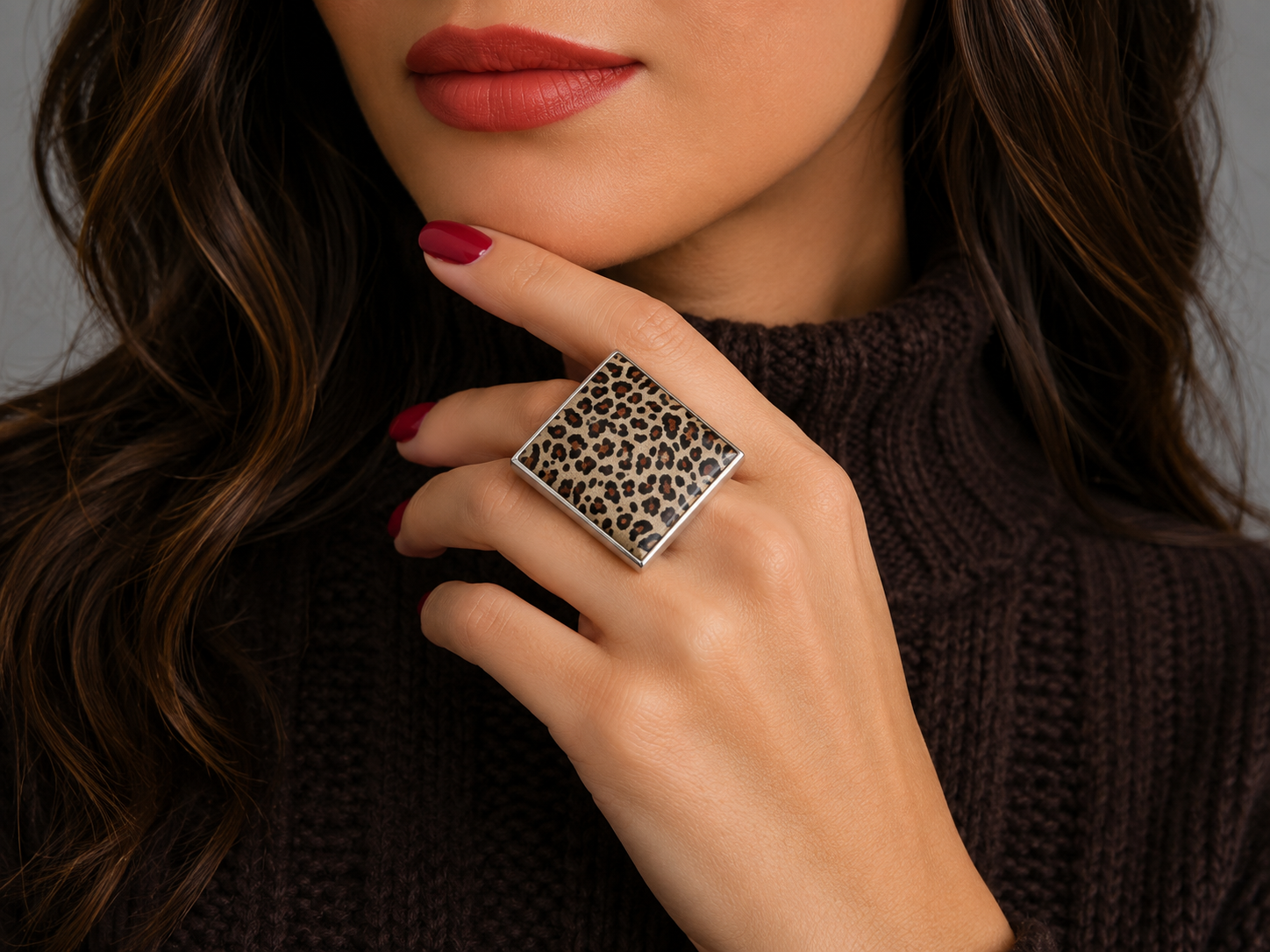 A close-up lifestyle shot of a woman wearing a large square silver ring with a leopard print pattern, styled with a dark brown knit sweater and red lipstick.