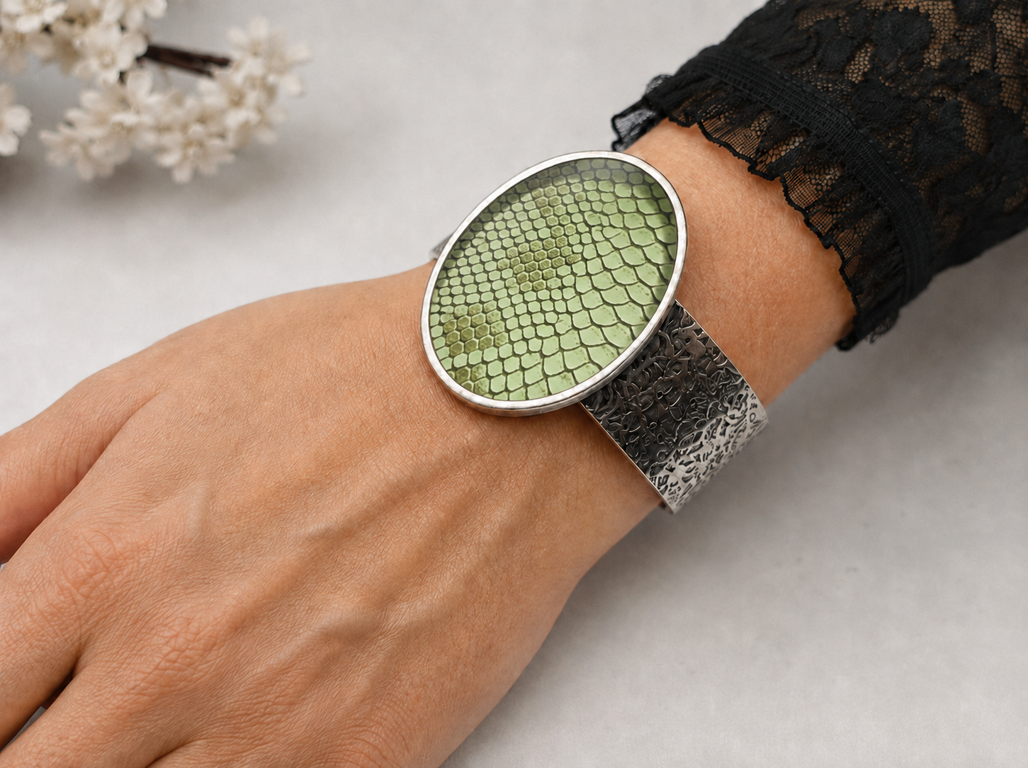 A close-up of a person's wrist wearing a silver-toned cuff watch with a textured green scale-pattern dial and a textured silver mesh band, styled with a black lace sleeve.