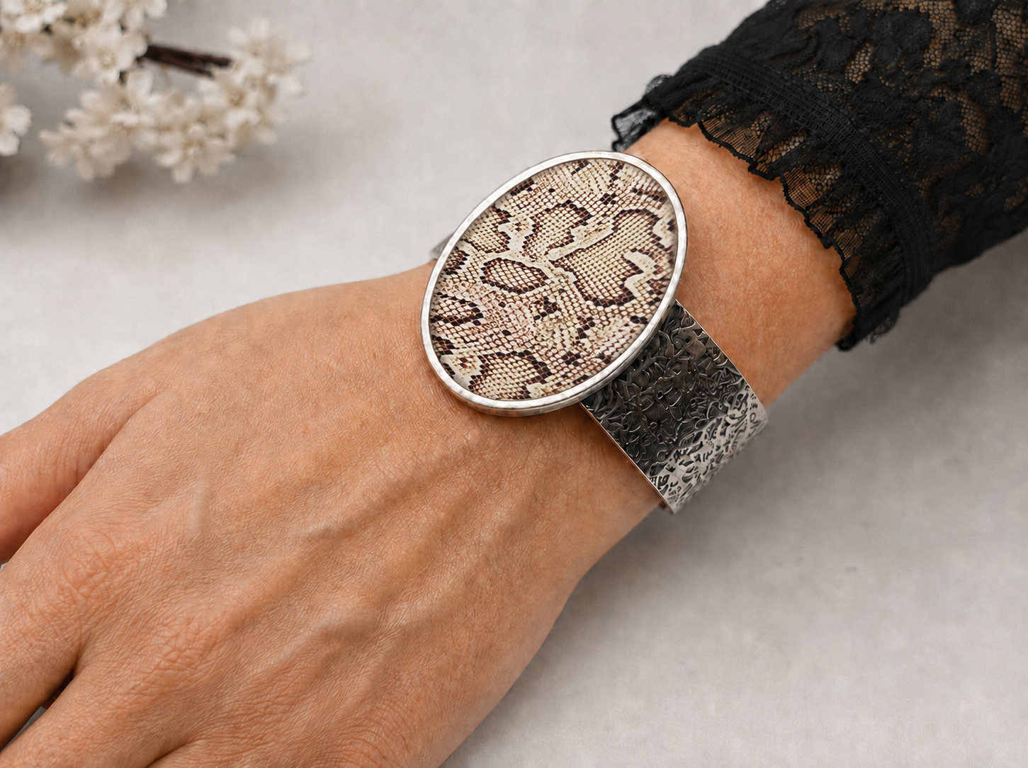 A close-up of a person's wrist wearing a wide silver cuff watch with an oval face featuring a beige and brown snakeskin pattern and a textured silver band.