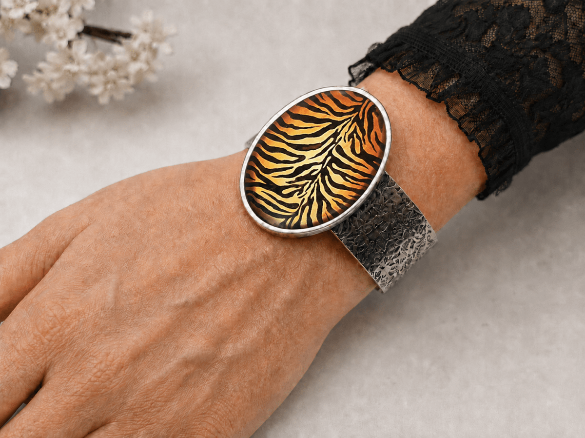 A close-up of a person's wrist wearing a wide silver cuff bracelet with an oval face featuring a yellow, orange, and black tiger-stripe pattern, paired with a textured silver mesh band.