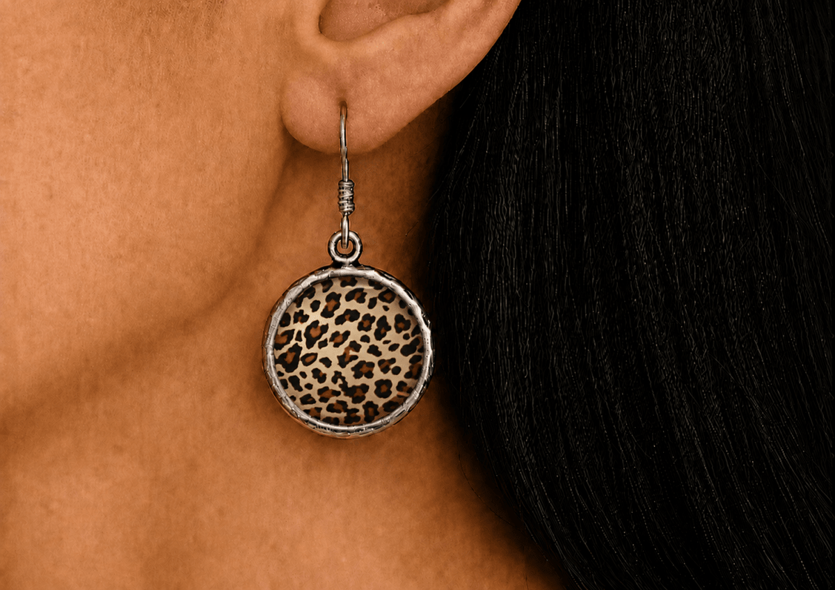 A close-up of a person's ear wearing a round dangle earring with a leopard print pattern and a silver-toned textured border.