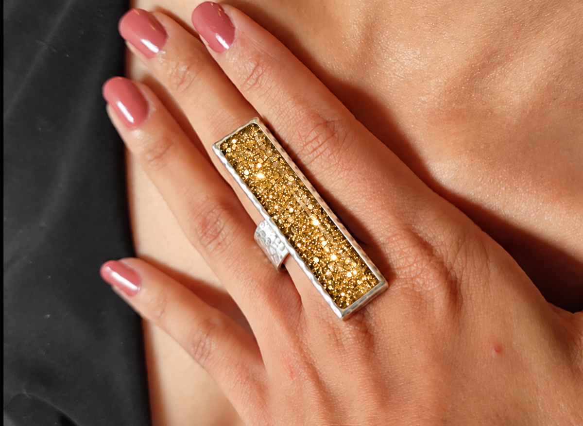 A close-up of a hand wearing a large rectangular silver ring with a sparkling gold glitter center.
