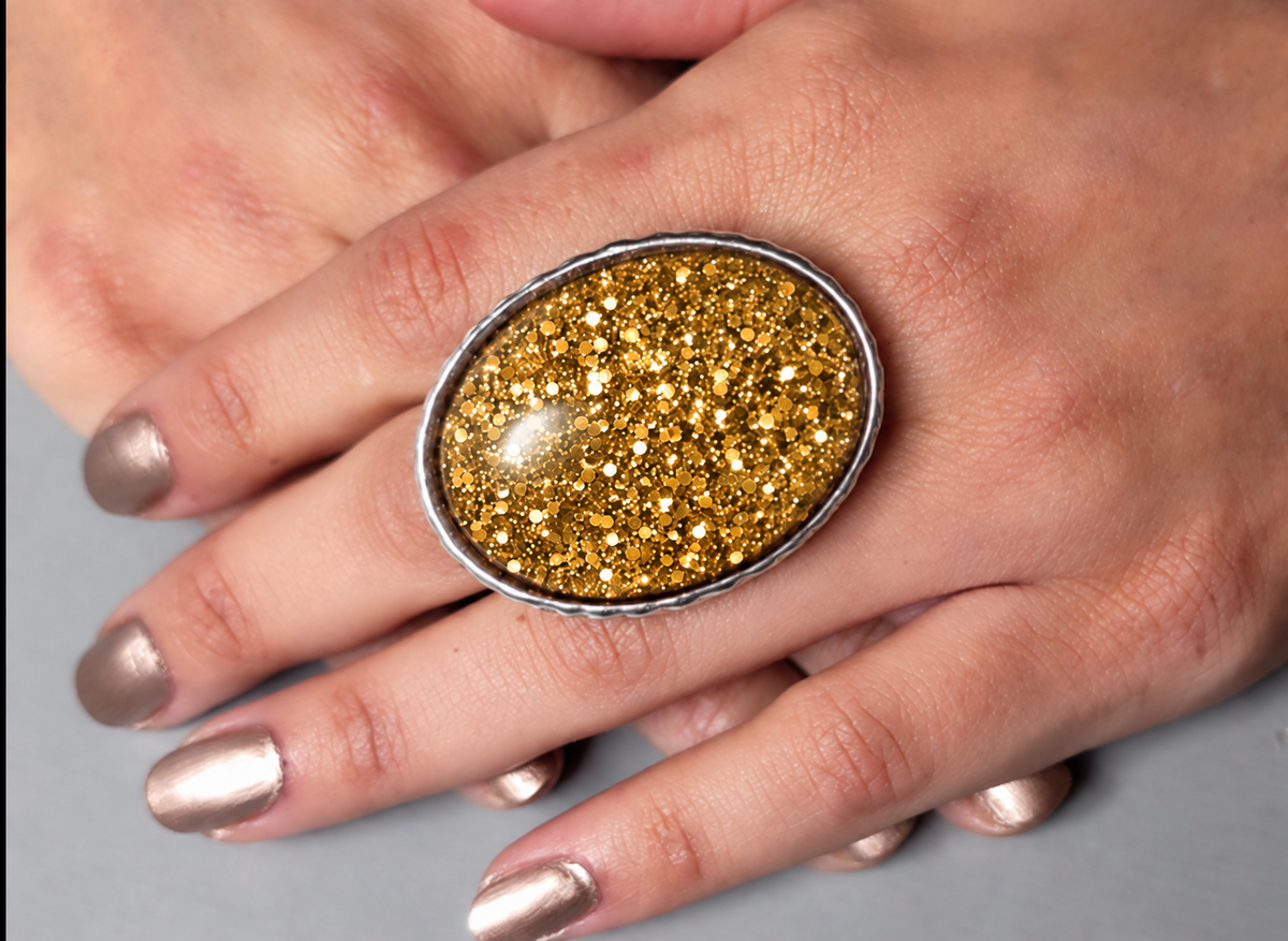 A close-up of a hand wearing a large oval ring with a textured, glittery gold center set in a silver-toned metal bezel.