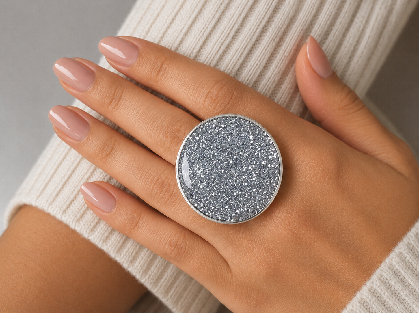 A close-up of a hand wearing a large round silver ring with a glittery gray-blue stone, styled with a cream ribbed knit sweater.