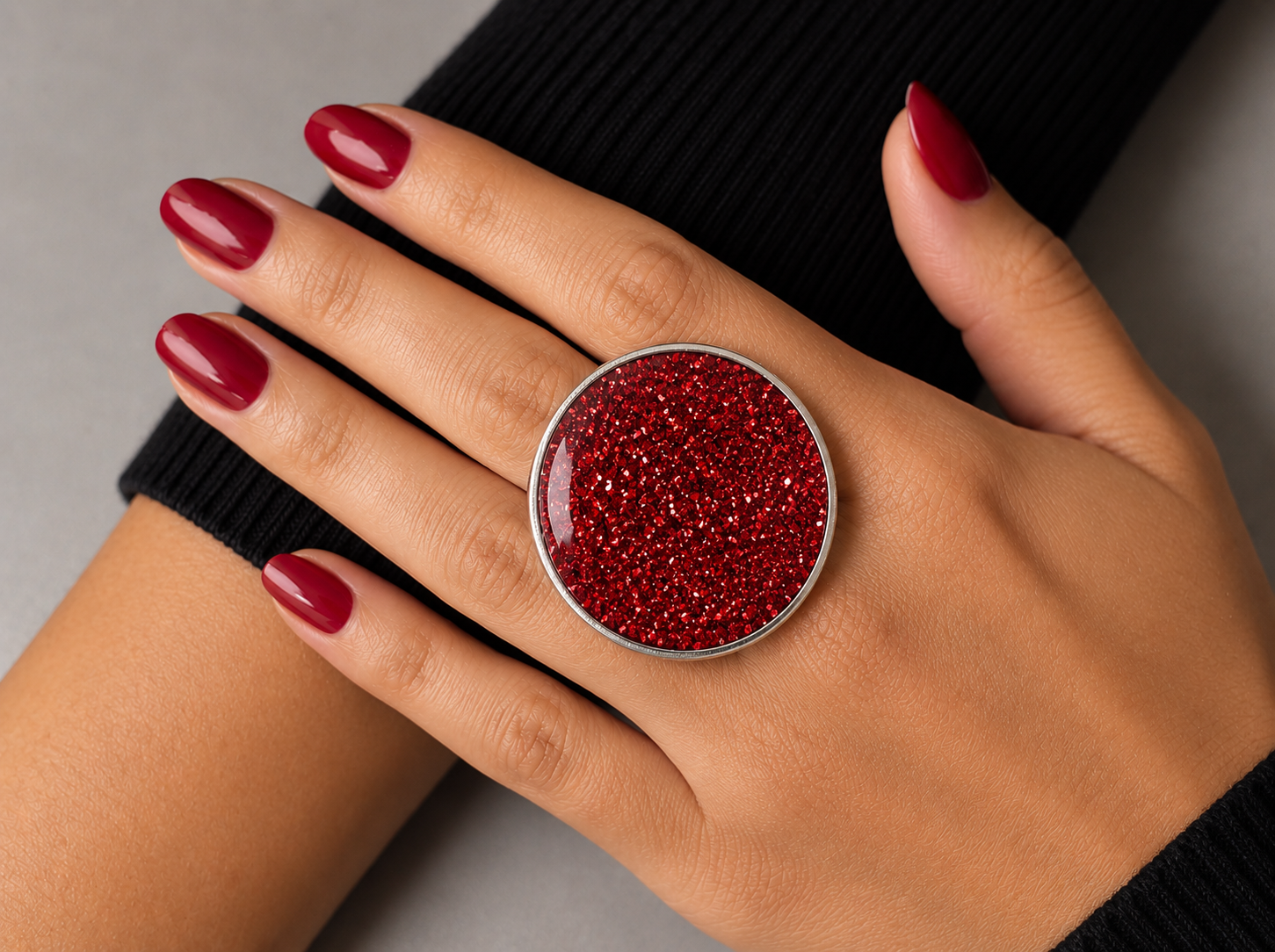 A close-up of a hand wearing a large round silver ring with sparkling red glitter, set against a black ribbed sweater and a neutral gray background.