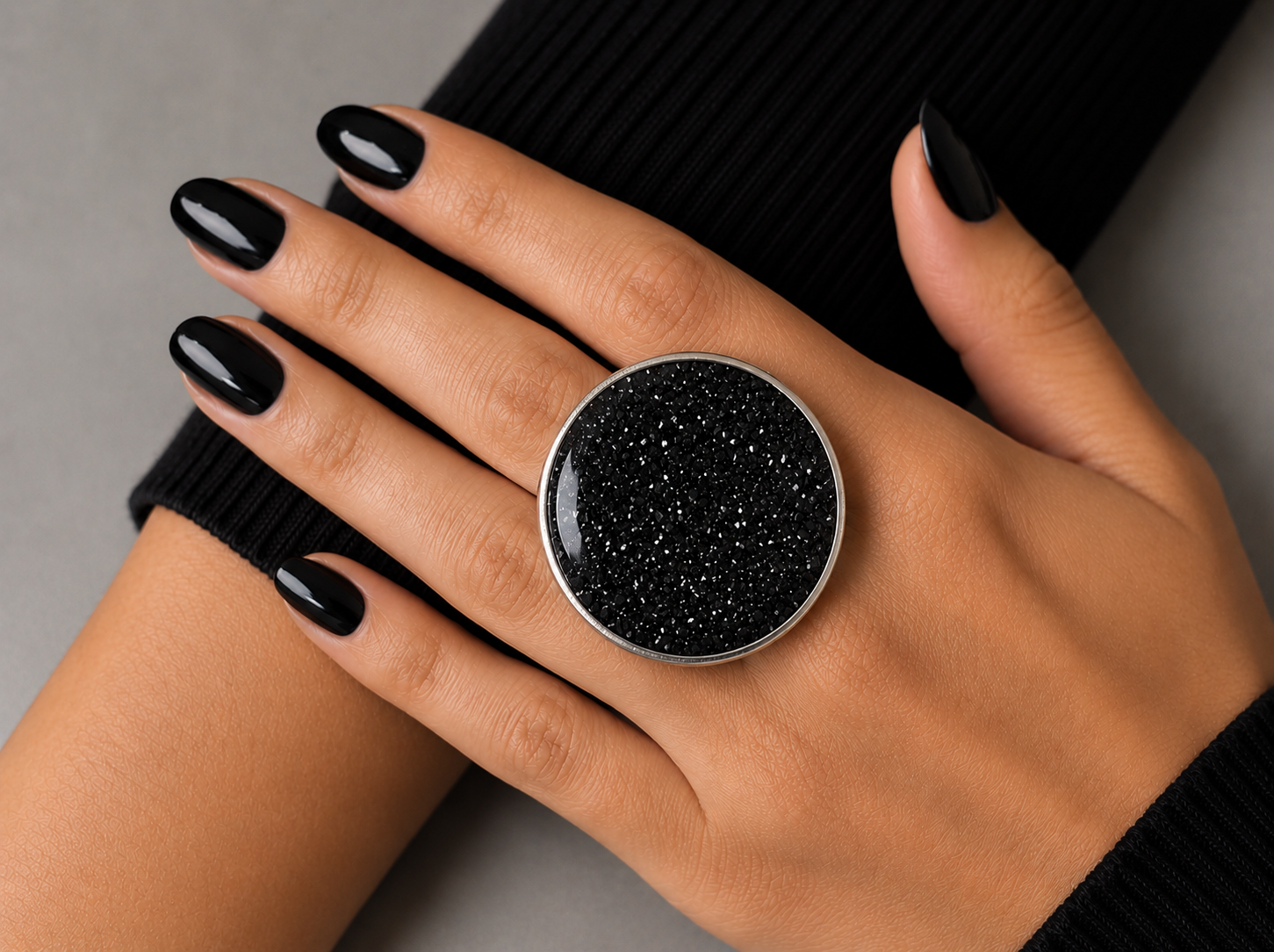 A close-up of a hand with black nail polish wearing a large round silver ring with a black glittery stone, resting against a black ribbed sleeve on a gray background.