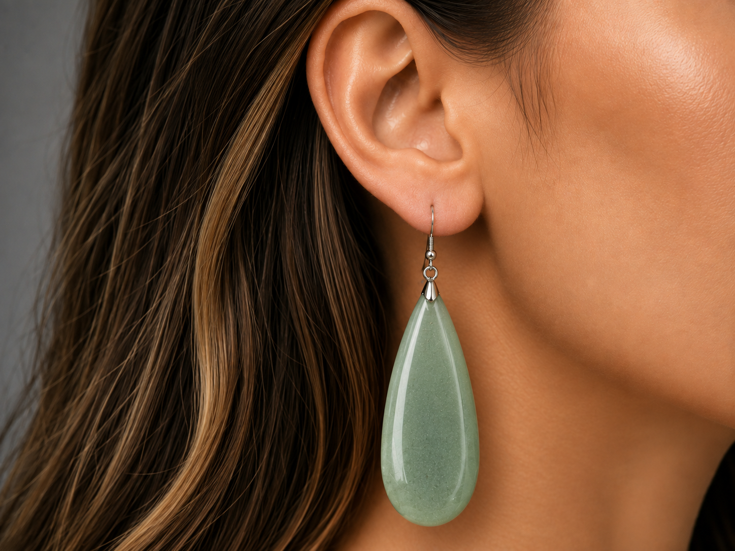 A close-up of a woman's ear wearing a large, polished light green teardrop-shaped dangle earring with a silver-toned fishhook back.