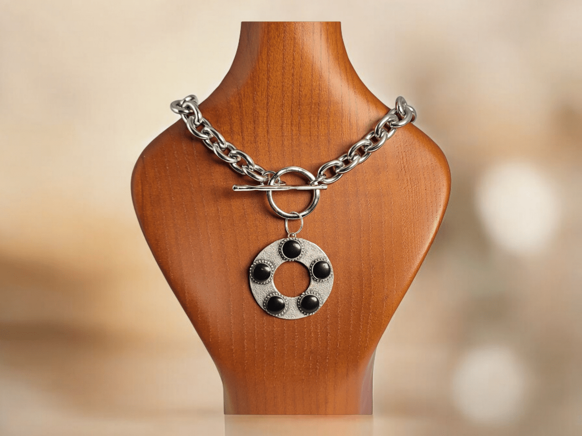 A large round silver and black pendant suspended from a chunky silver chain on a wooden bust Main image
