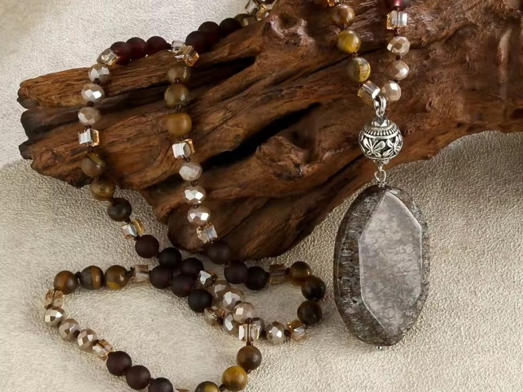 A beaded necklace with a large polished gray stone pendant, displayed on a piece of driftwood over a neutral fabric background. The necklace features faceted brown, smoky clear, and dark brown beads with a silver-toned filigree accent.
