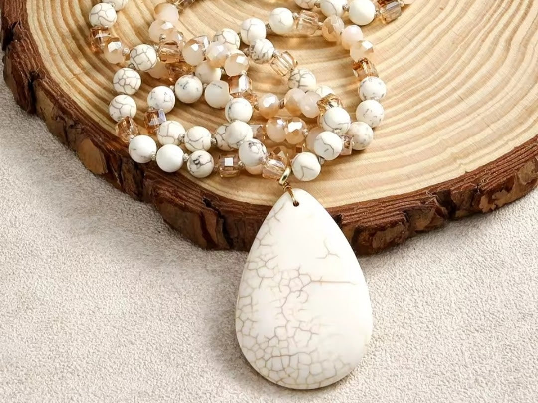 A beaded necklace with white and cream marbled beads and a large white teardrop pendant with a crackled pattern, displayed on a rustic wood slice over a light beige fabric background.