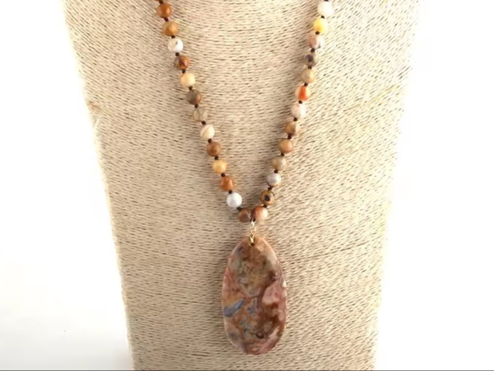 A beaded necklace with a large teardrop-shaped polished stone pendant, displayed on a beige fabric jewelry bust. The necklace features small marbled beads in earthy brown, tan, and cream tones, with a large brown, pink, and blue marbled pendant.