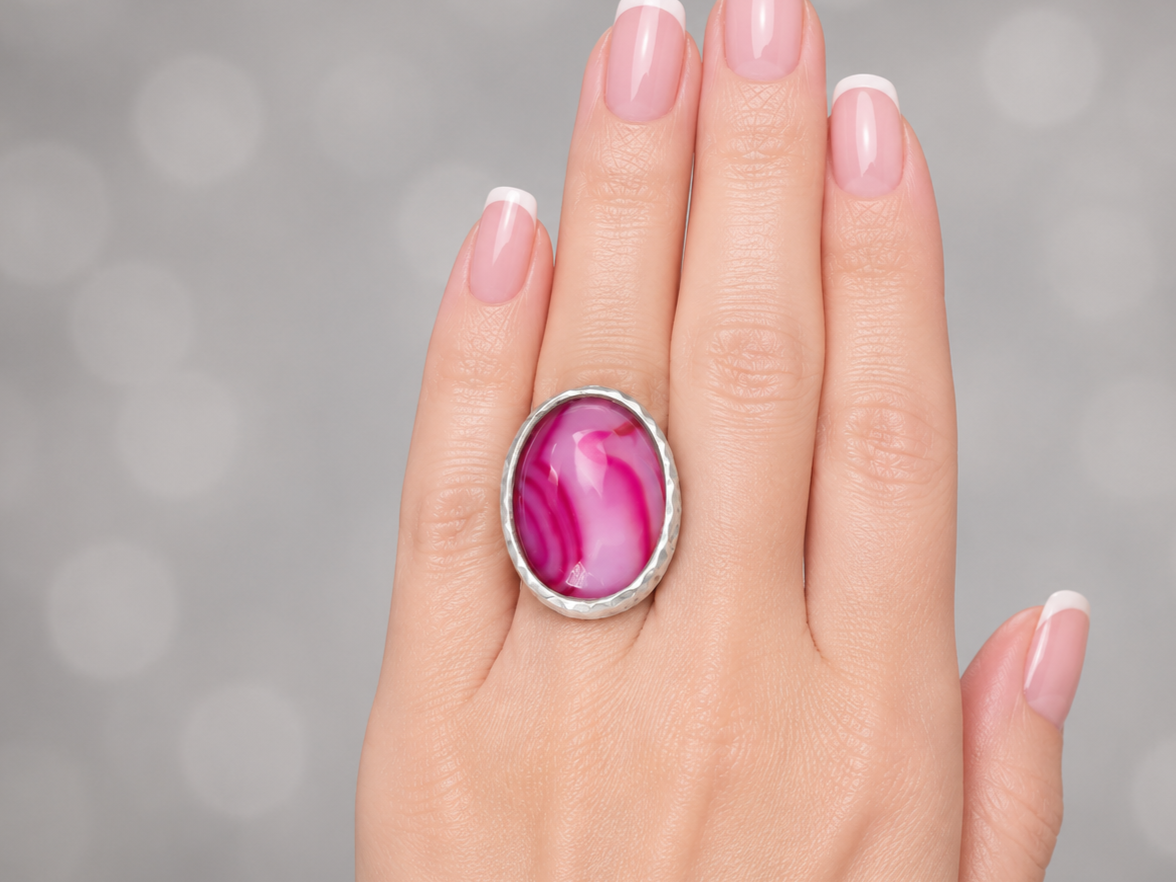 A close-up of a hand wearing a silver ring with an oval pink agate stone, set against a soft gray bokeh background.