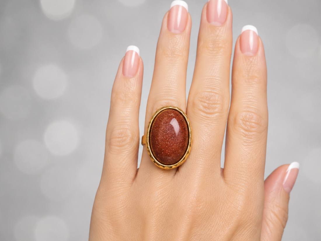 A gold ring with a large oval reddish-brown stone, worn on a hand with a French manicure against a soft gray bokeh background.