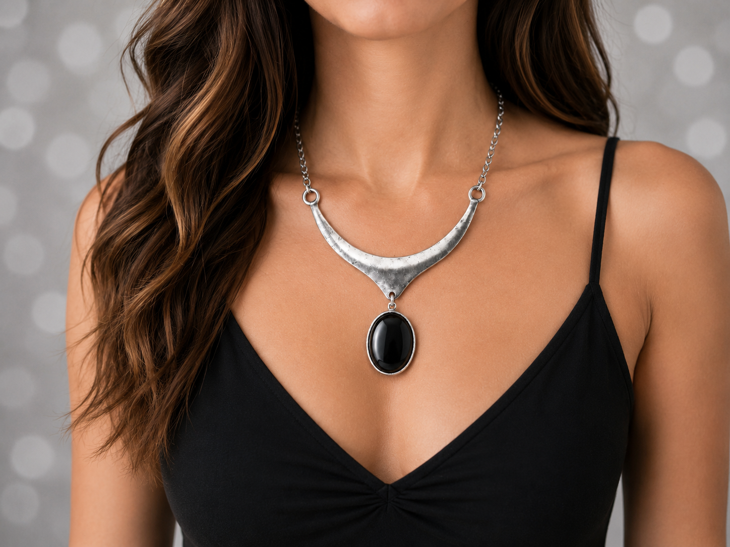 A close-up of a woman wearing a silver crescent-shaped statement necklace with a black oval pendant, paired with a black spaghetti-strap top against a soft gray bokeh background. Main image