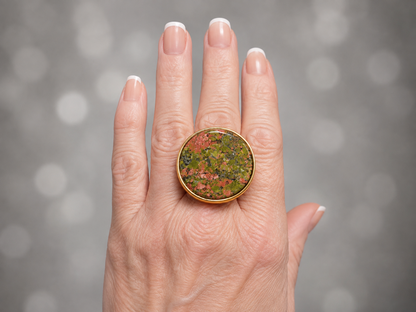 A close-up of a hand wearing a large round gold ring with a textured, marbled green and pink stone, set against a soft gray bokeh background.