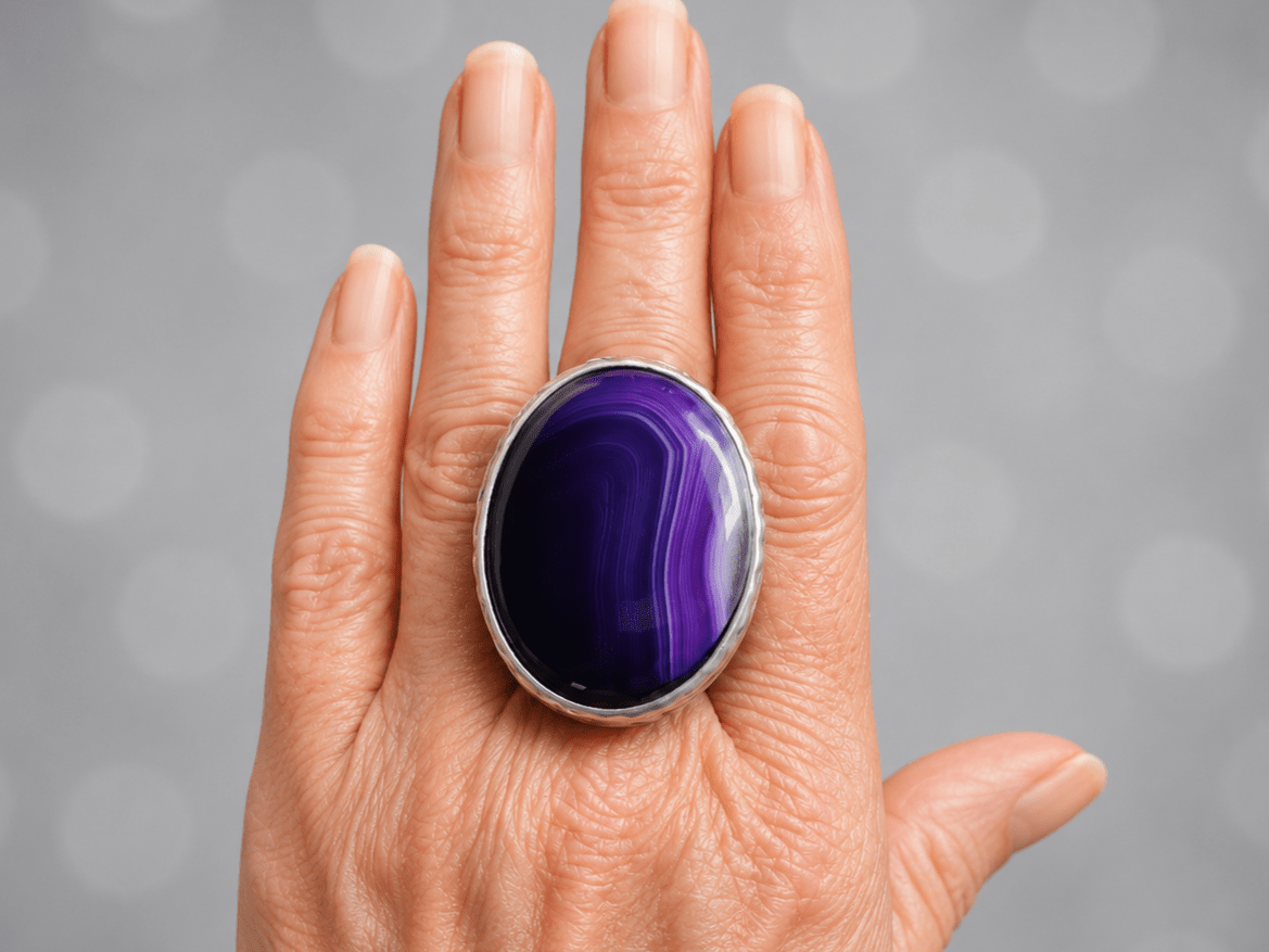 A close-up of a hand wearing a large oval ring with a polished purple agate stone in a silver bezel setting, against a soft gray bokeh background.