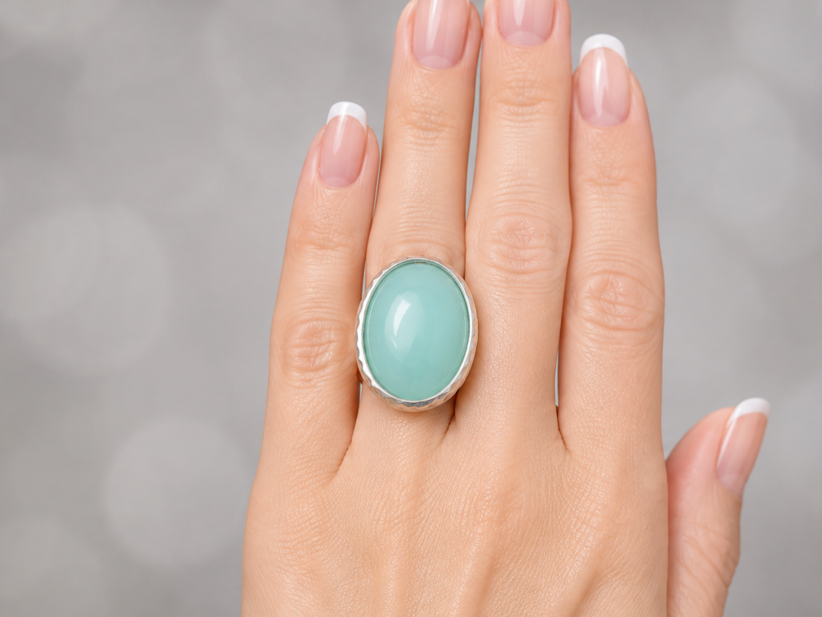 A silver ring with a large oval light green cabochon gemstone, shown worn on a hand with a French manicure against a soft gray bokeh background.