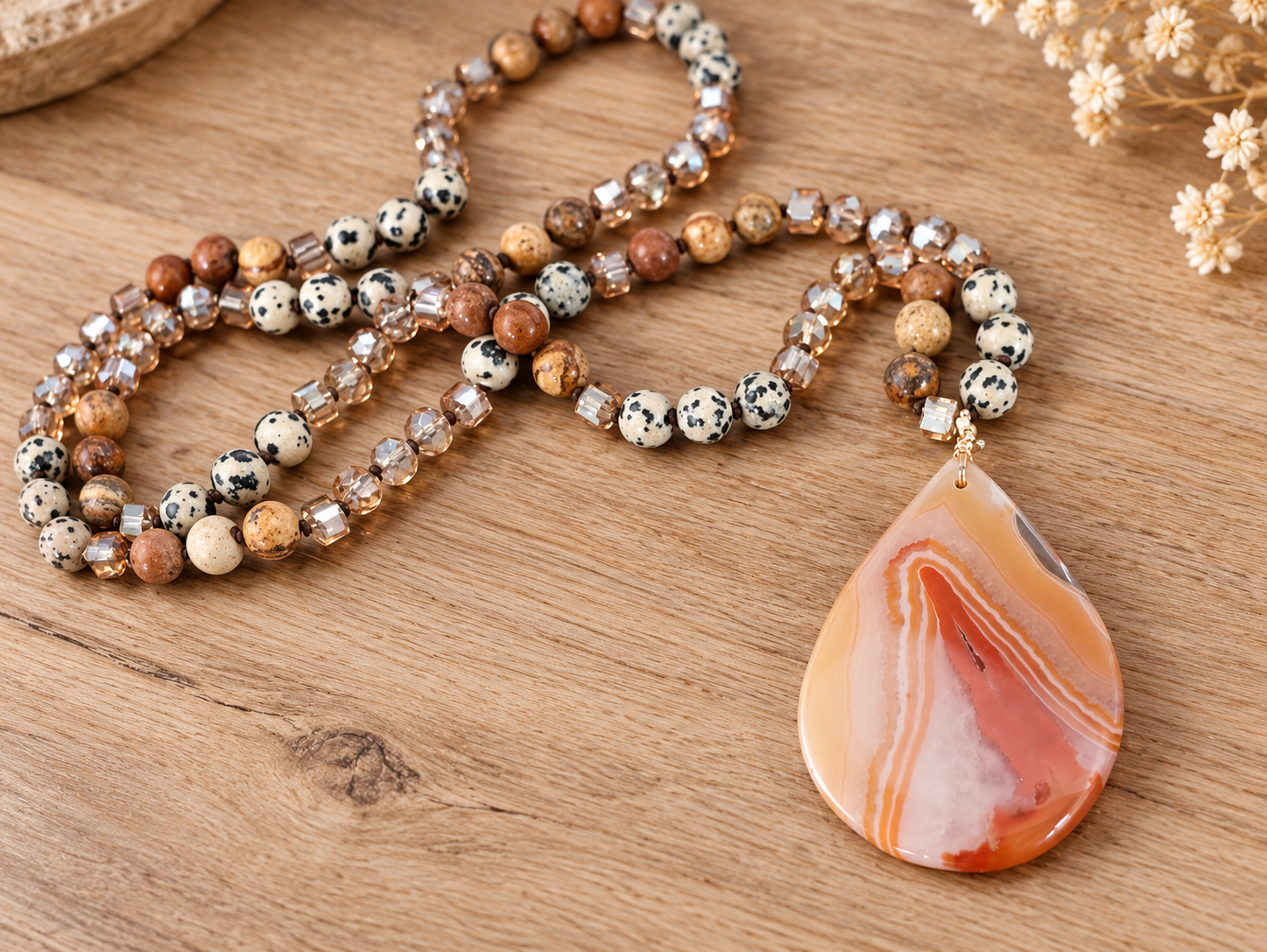 A beaded necklace with a large teardrop pendant featuring orange, white, and red agate-like banding, displayed on a wooden surface alongside brown and speckled beads.