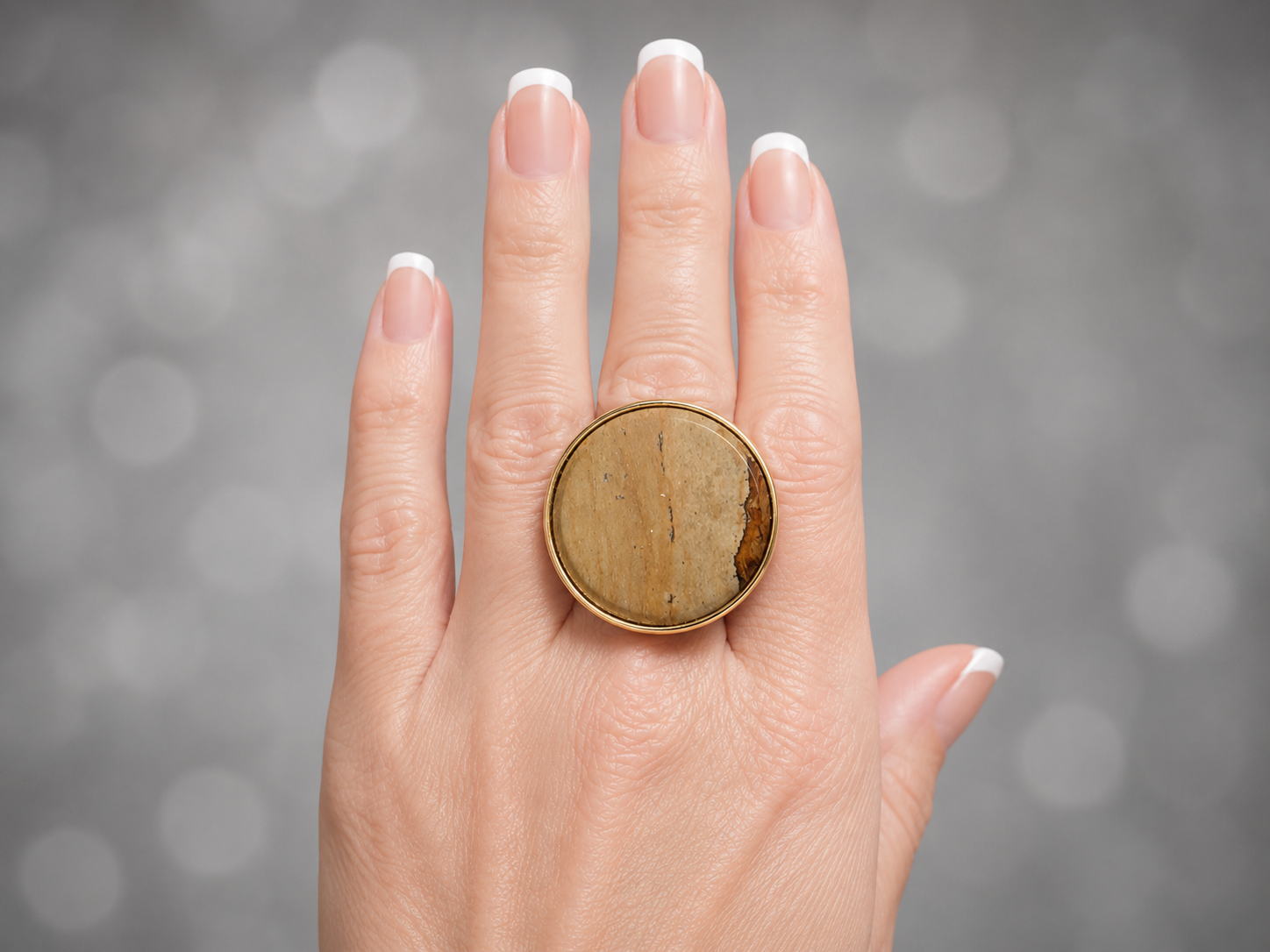 a close up of a models hand against a bokheh background. she is wearing a picture jasper gold ring