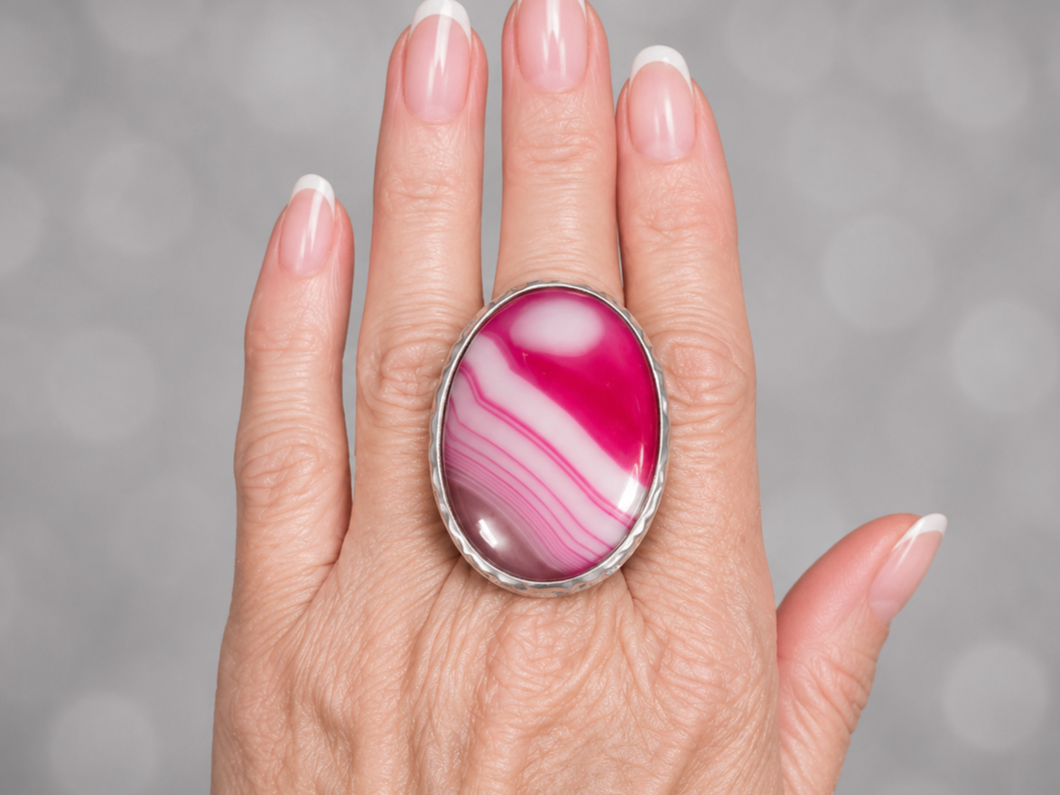 A close-up of a hand wearing a large oval ring with a pink and white banded agate-style stone in a silver bezel setting, against a soft gray bokeh background.