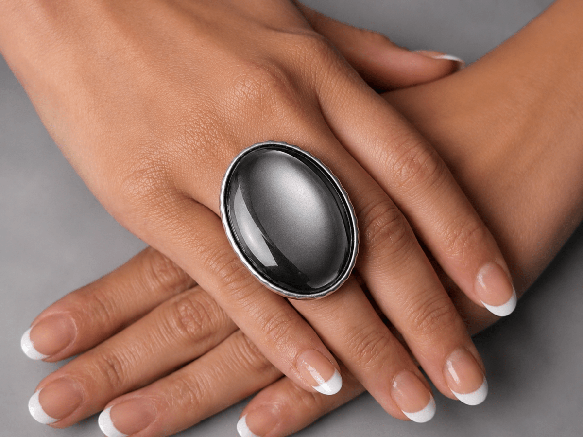 A close-up of a person's hands wearing a large oval black cabochon ring with a silver-toned metal border, set against a neutral gray background. Main image